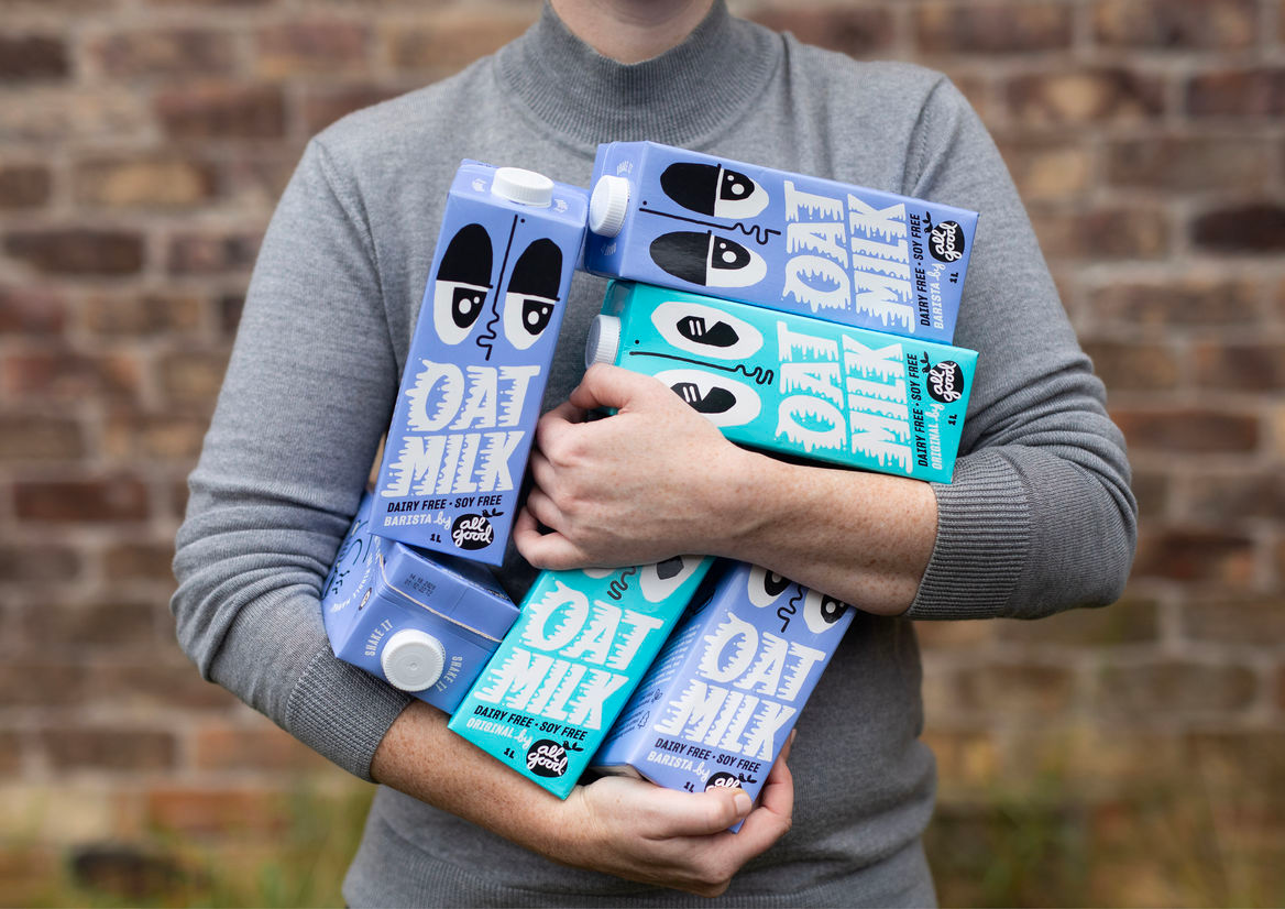 A person holding a stack of oat milk cartons from the All Good brand, a company focused on Fairtrade and organic products