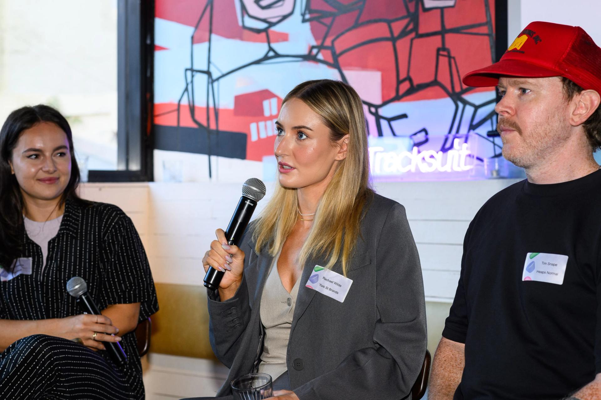 Rachael Wilde, Co-Founder & CMO at tbh Skincare, holding a microphone and discussing marketing strategies during a panel discussion at a Tracksuit event, sitting between two male panelists