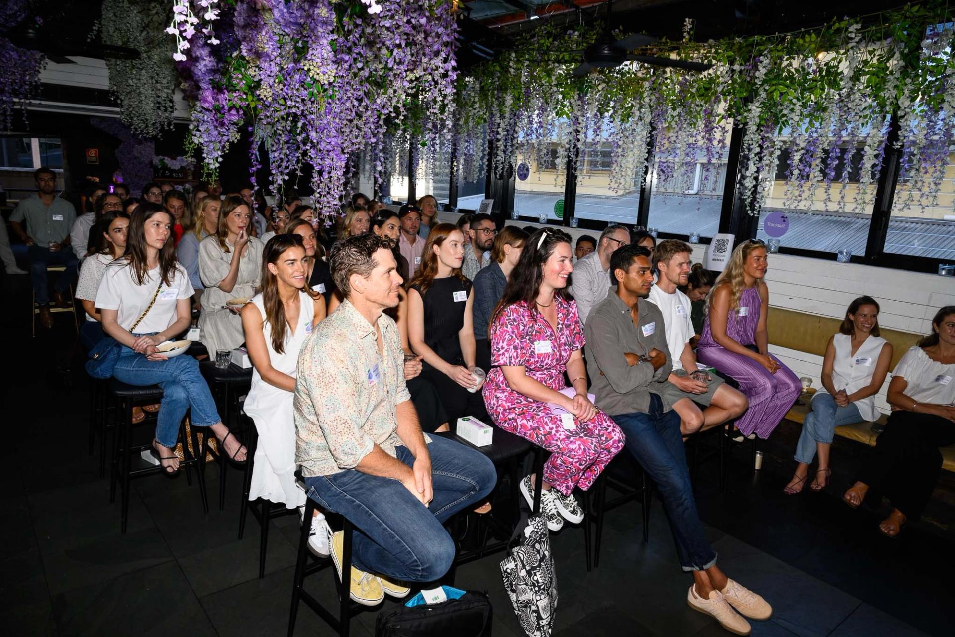 A large group of marketing professionals gathered in a room for a Tracksuit community event, with purple flowers hanging from the ceiling for decoration.