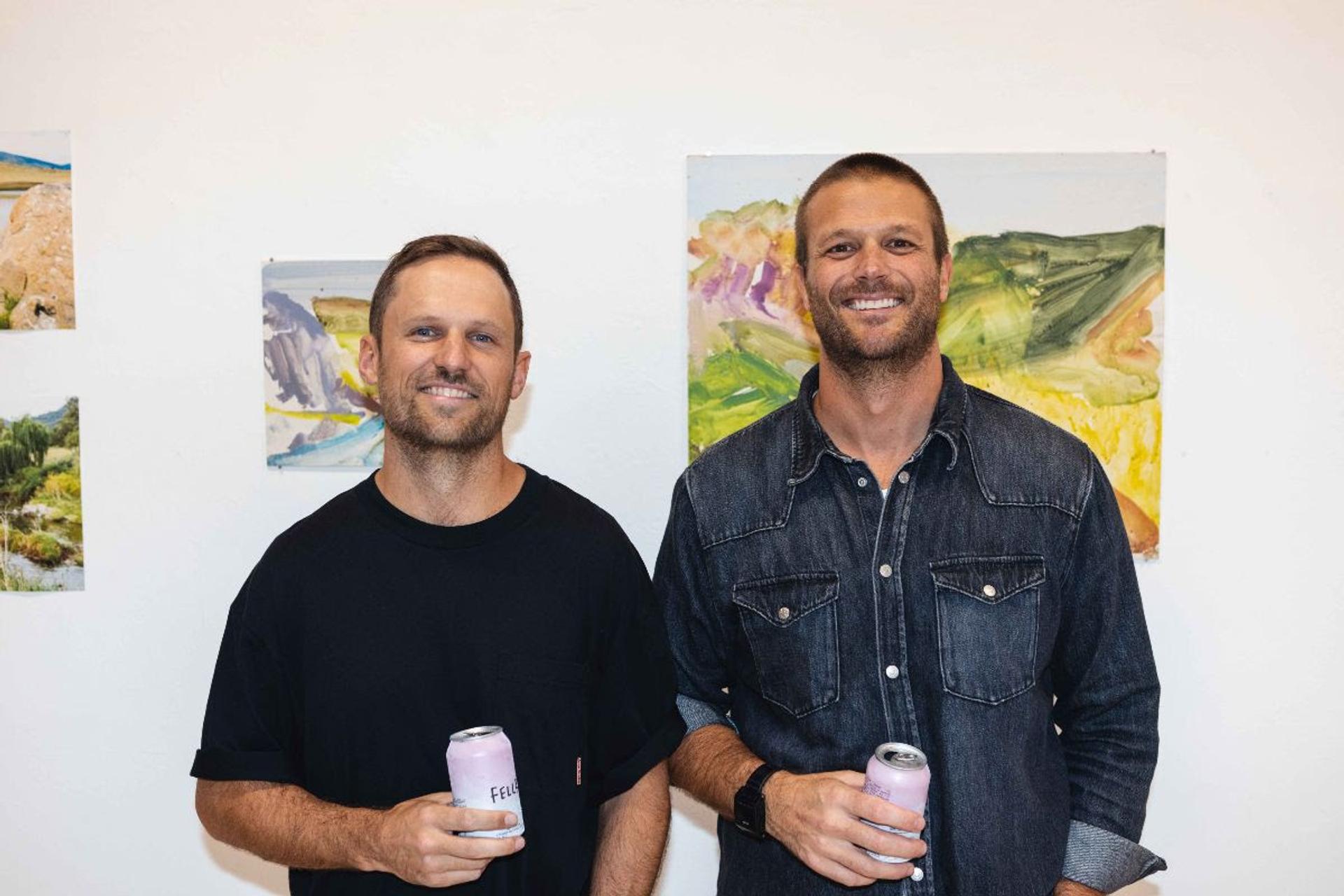 Co-founders of FELLR, Andy Skora and Will Morgan, holding cans of their popular alcoholic seltzer in front of a painting, symbolizing their creative journey in the beverage industry.