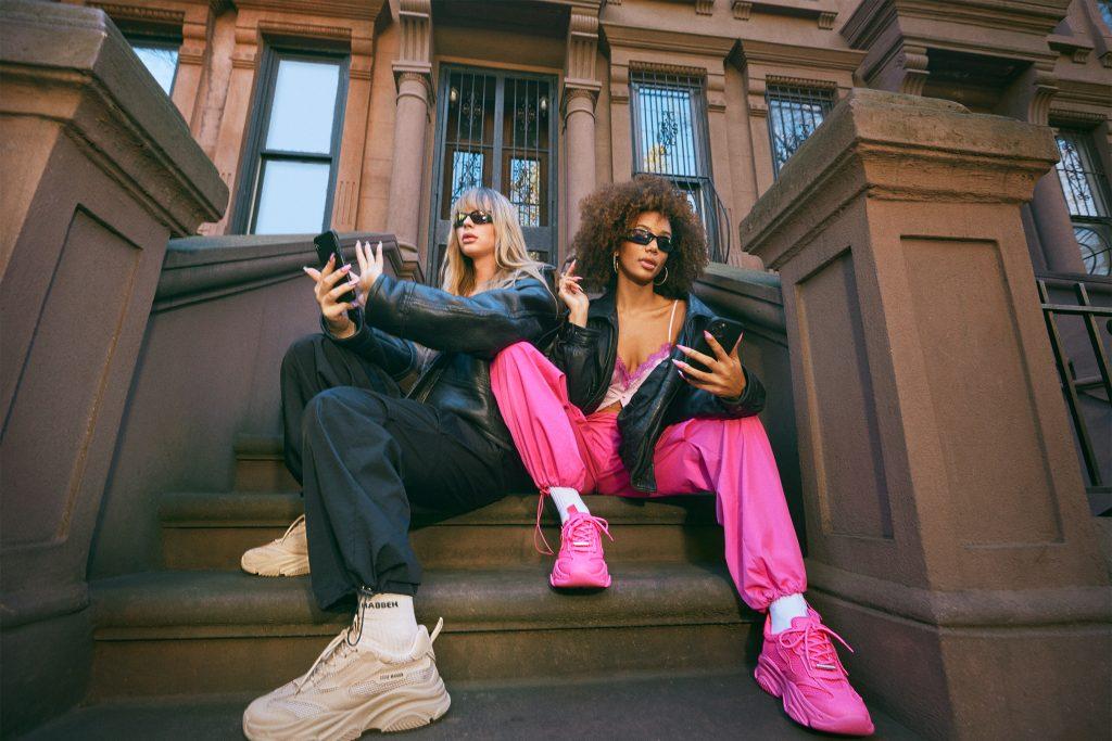 Two women engrossed in their mobile phones while sitting on the steps of a building, representing the modern, tech-savvy audience of Steve Madden footwear brand