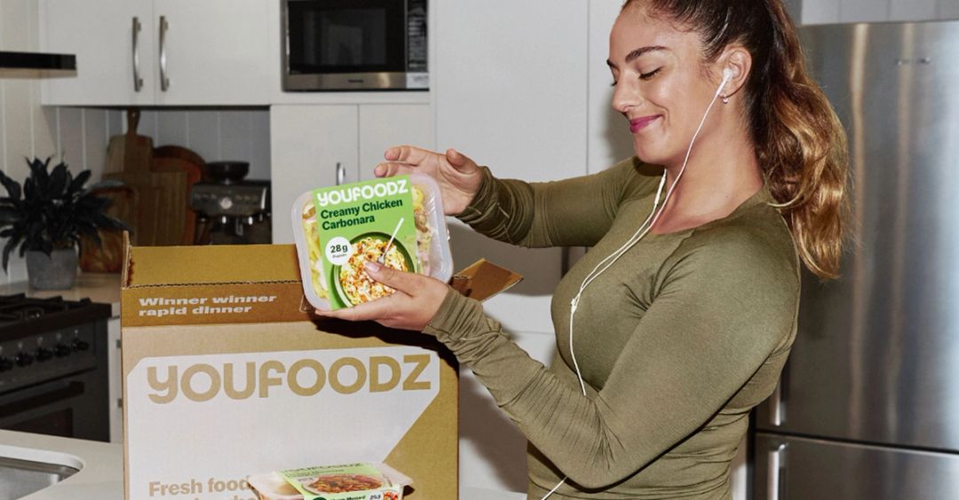 A smiling woman in an olive long-sleeve top and earphones holds up a Youfoodz Creamy Chicken Carbonara meal above an open Youfoodz delivery box in a white kitchen.
