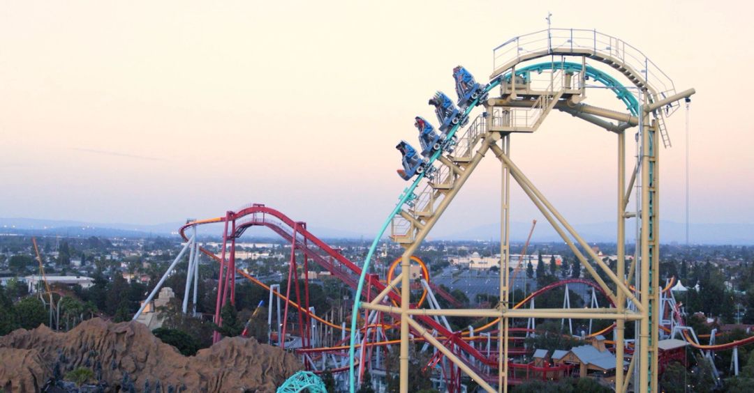 A roller coaster train climbing a tall cream-coloured track at dusk, with red and teal coaster tracks and a theme park below.
