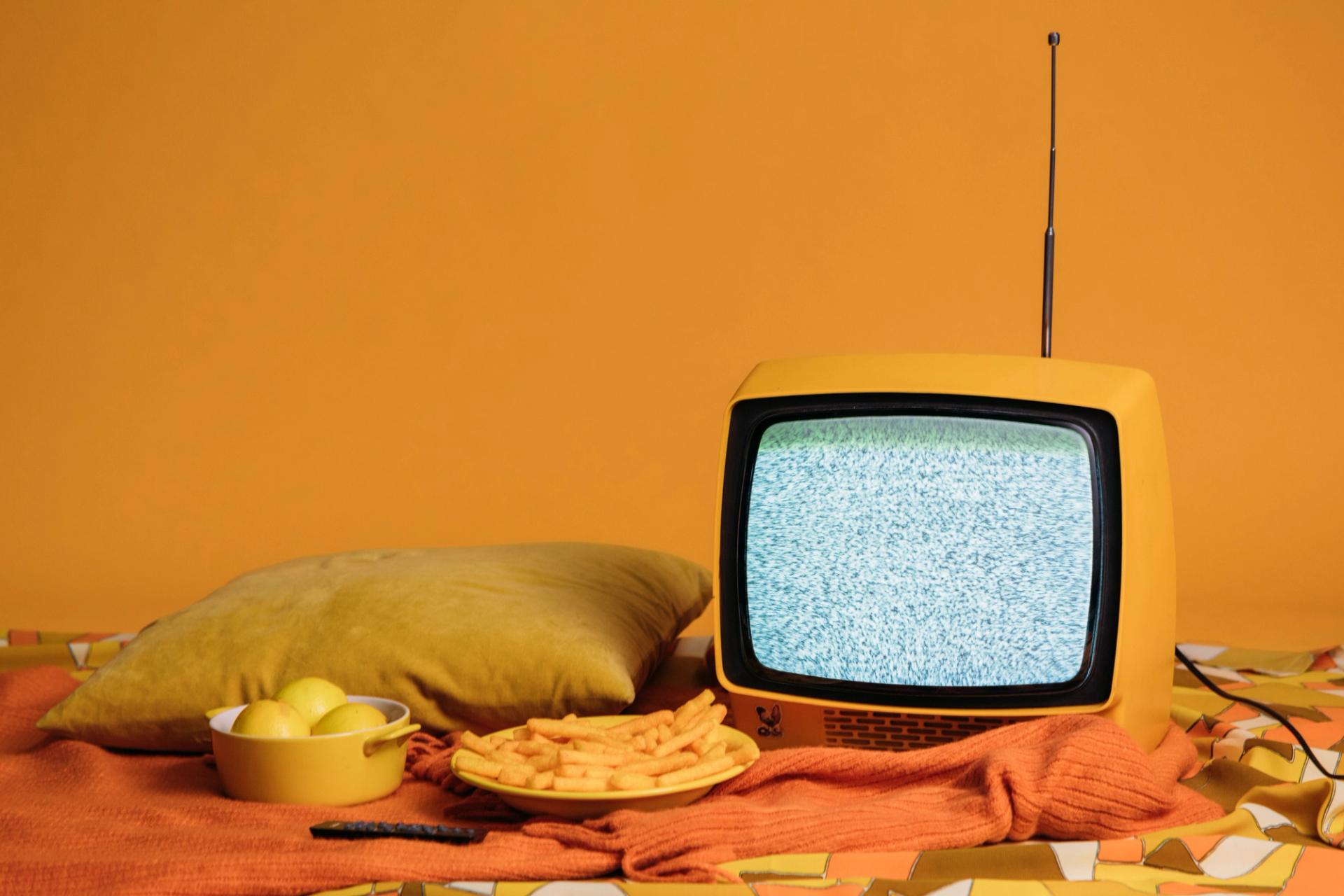 A vibrant yellow television on a bed displaying a TV advertising related content, accompanied by a bowl of fresh lemons and a plate of delicious french fries, symbolizing the blend of traditional and digital media consumption.
