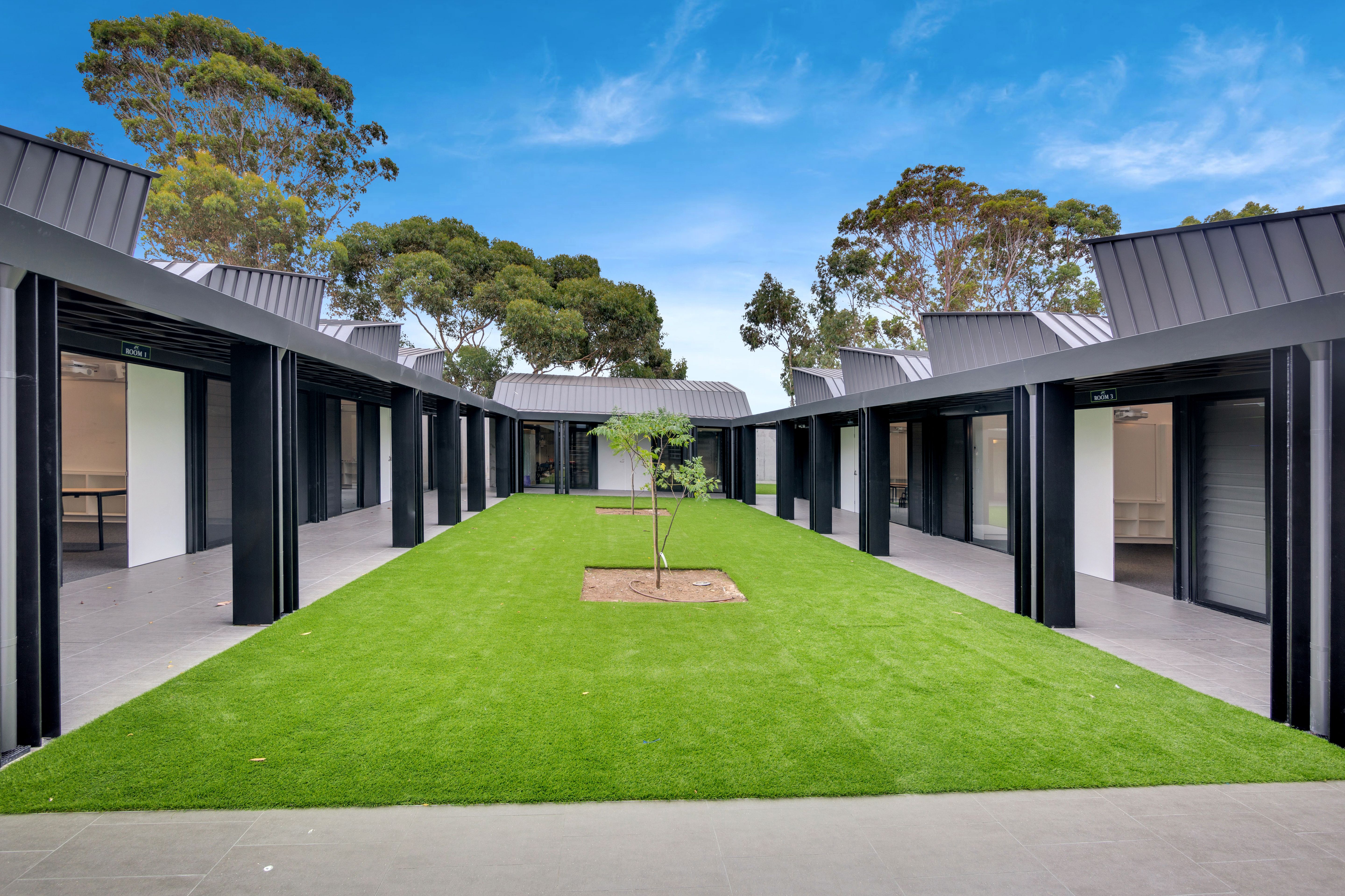 Modern building complex with dark roofs and covered walkways surrounding a central green courtyard with a young tree.