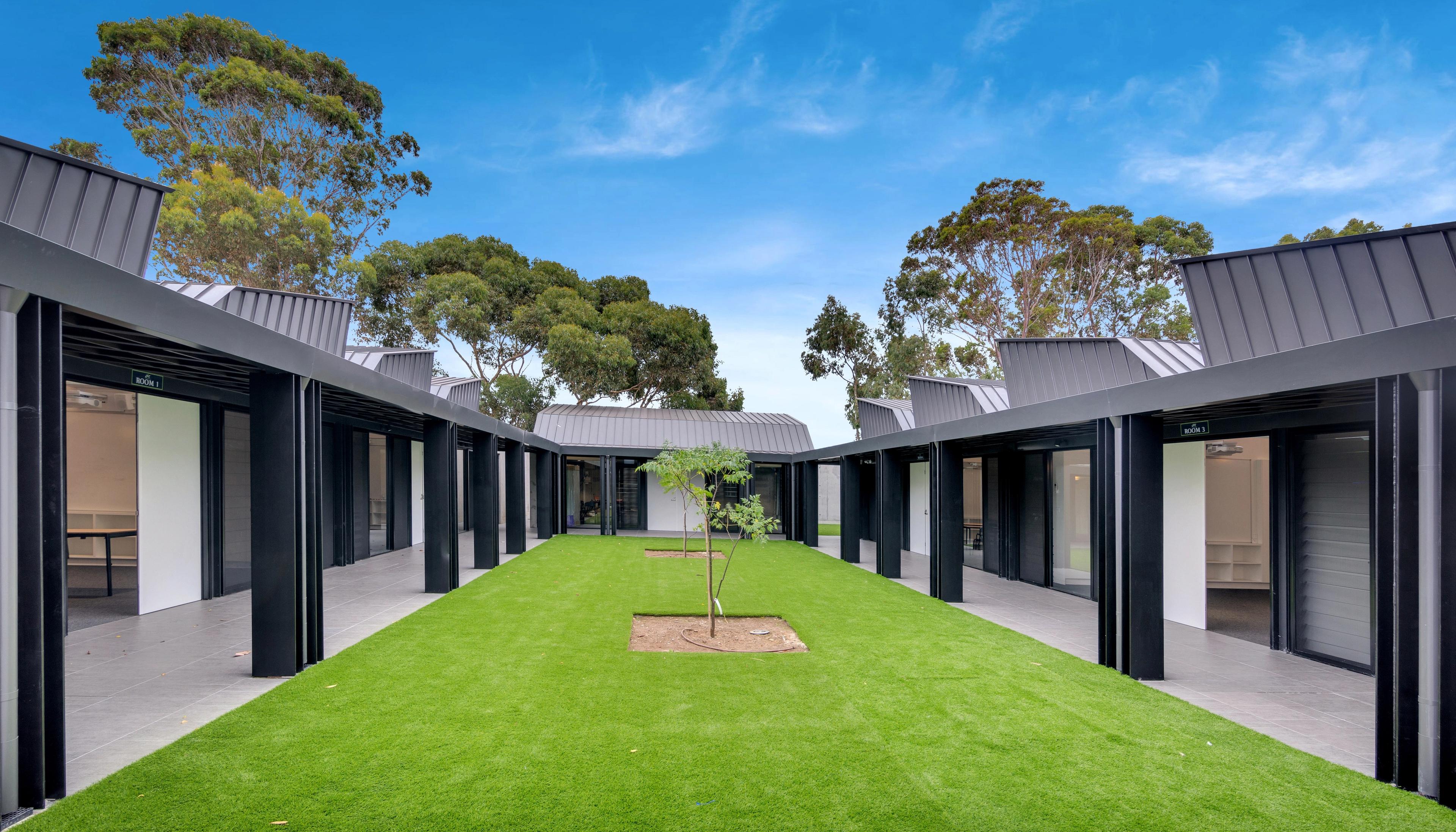 Modern building complex with dark roofs and covered walkways surrounding a central green courtyard with a young tree.