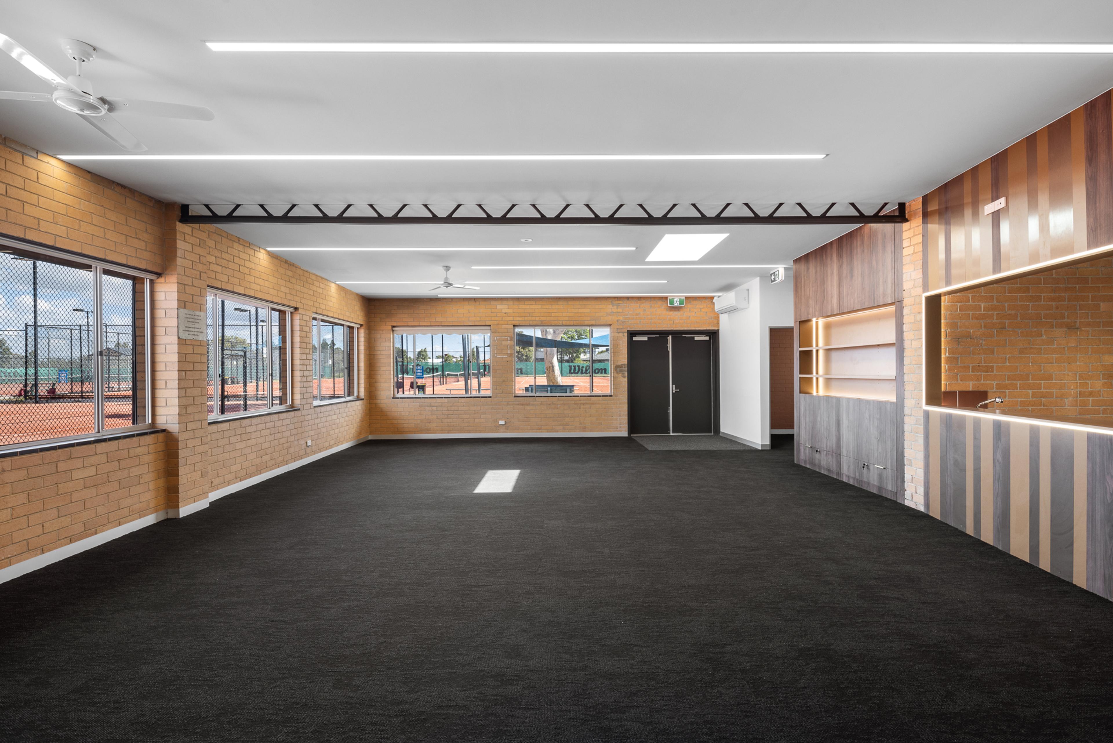 Empty modern clubroom with dark carpet, brick walls overlooking tennis courts, and a wood-paneled counter.