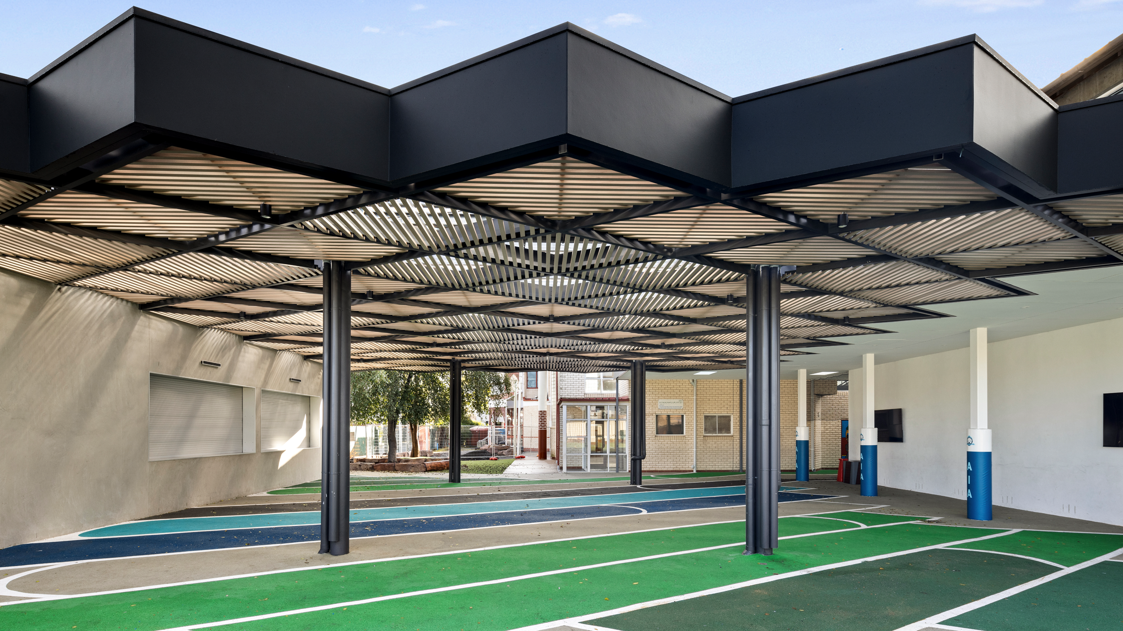 A modern outdoor area featuring a dark, slatted geometric canopy covering a colorful paved court with white lines.
