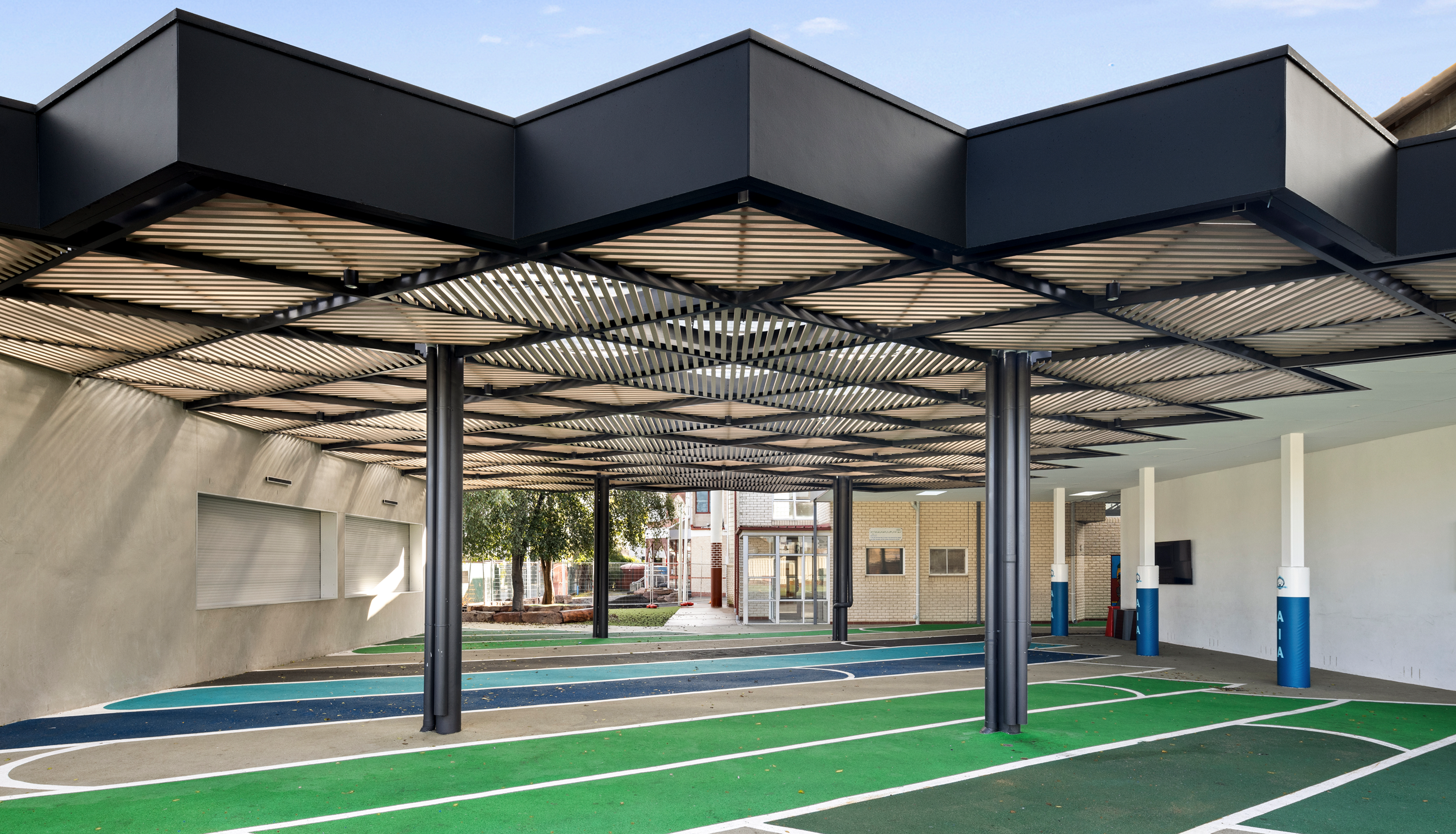 A modern outdoor area featuring a dark, slatted geometric canopy covering a colorful paved court with white lines.