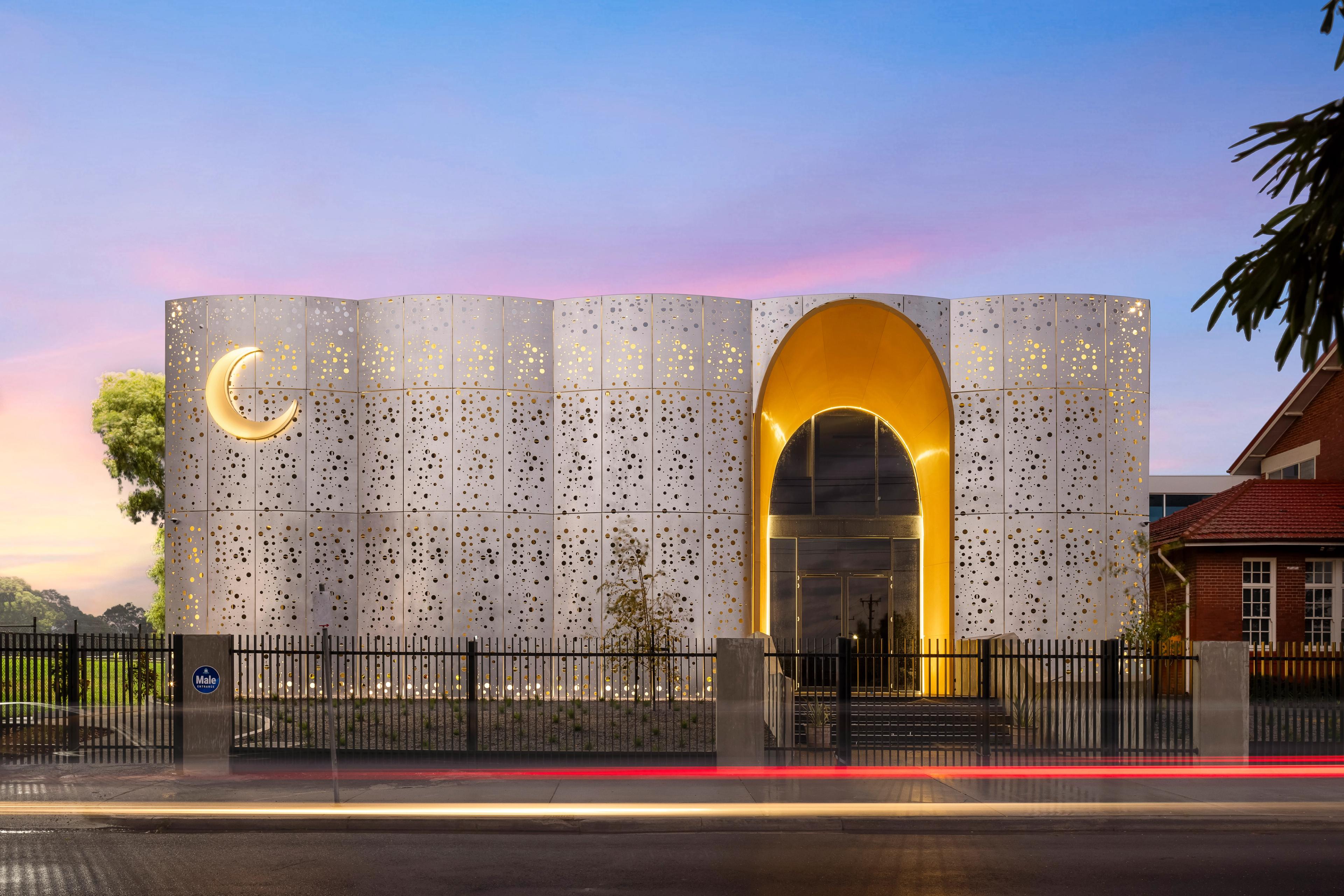 Modern Islamic center with a white perforated facade, golden crescent moon, and glowing arched entrance at twilight.