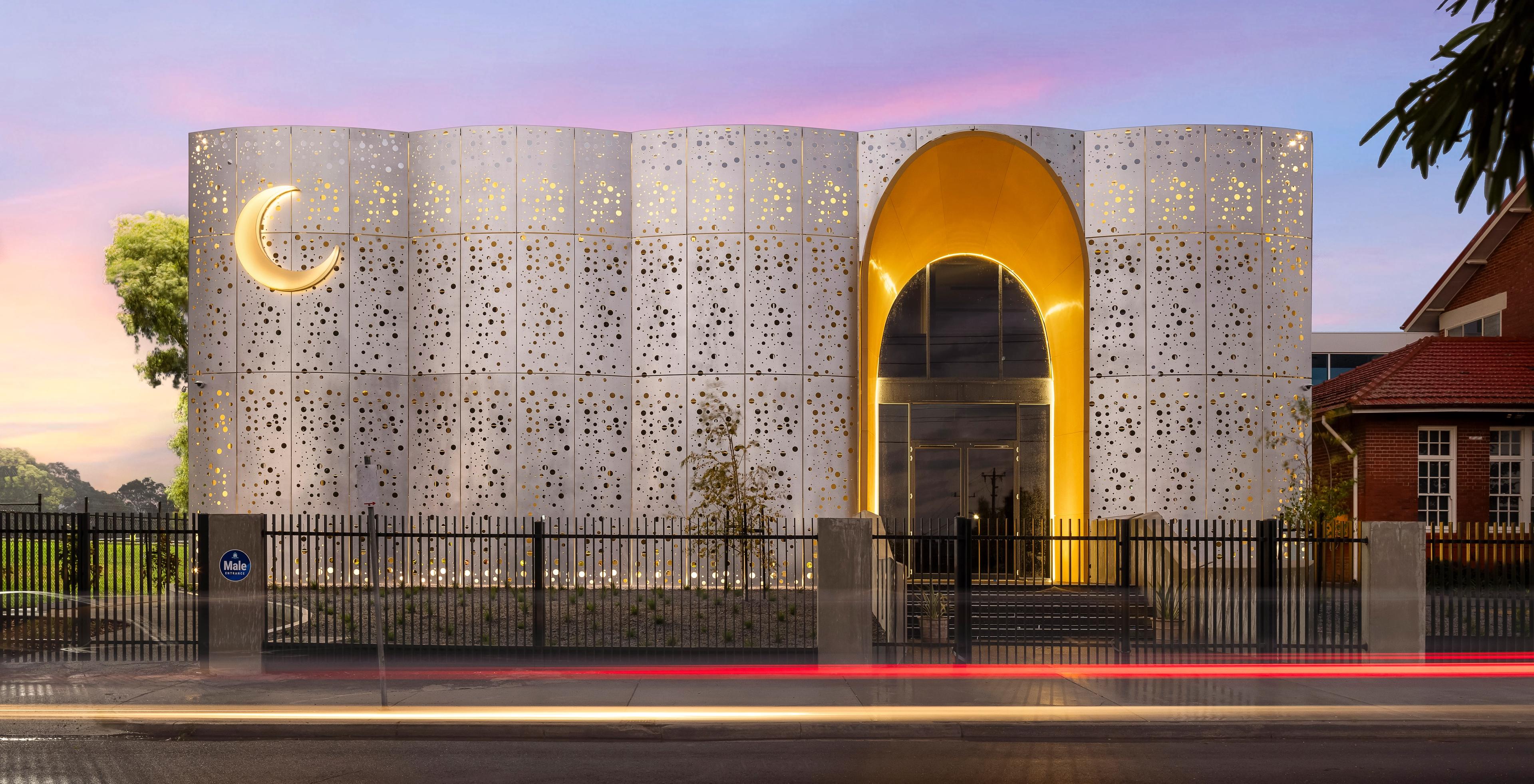 Modern mosque with a perforated white facade, a golden crescent moon, and an illuminated arched entrance at dusk.