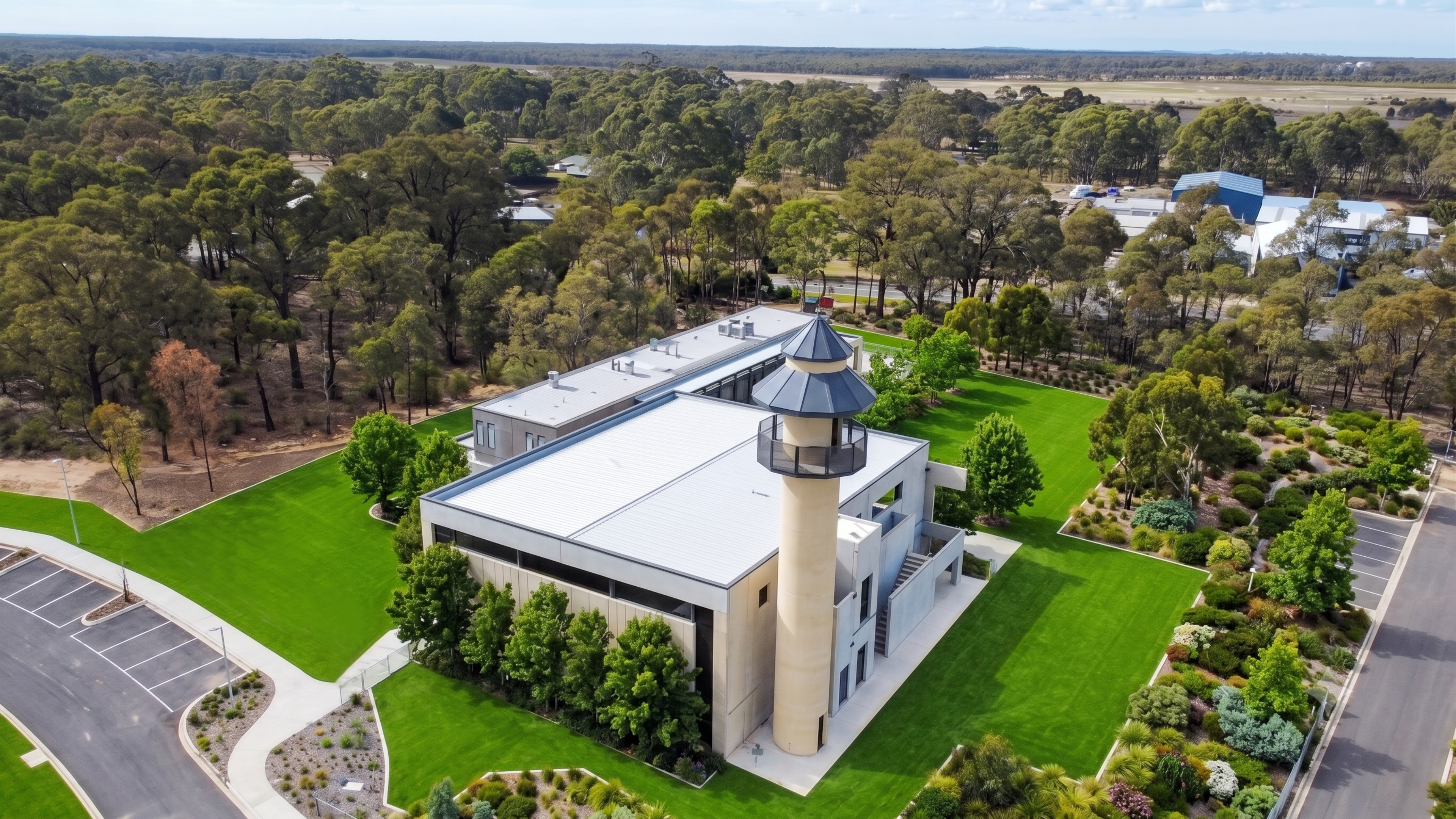 Aerial view of a modern building with a tall, cylindrical tower, surrounded by lush green lawns and landscaped gardens.