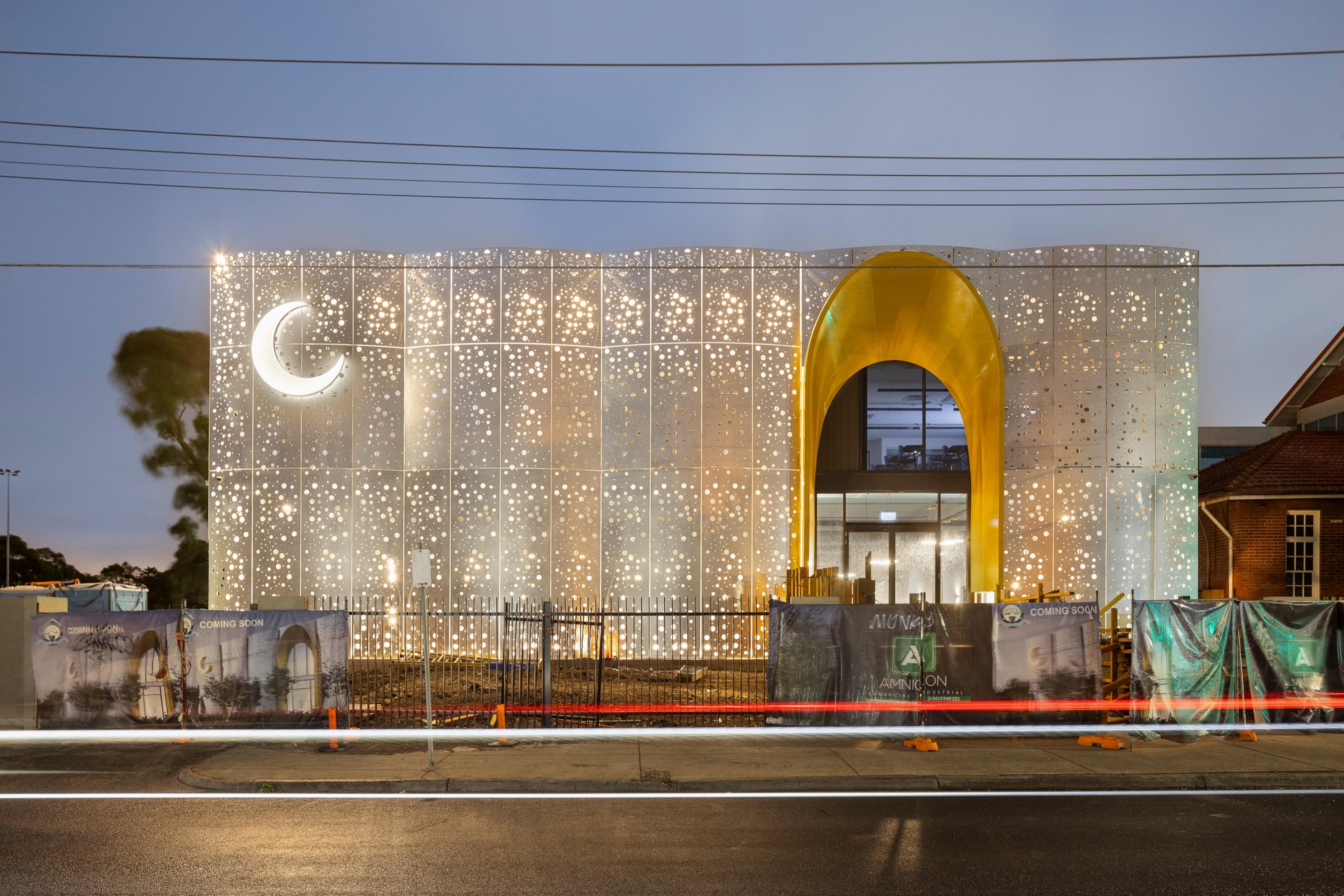 Modern building at night with a glowing, star-patterned facade, a large crescent moon symbol, and a golden arched entrance. Construction site barriers and light trails are in the foreground.