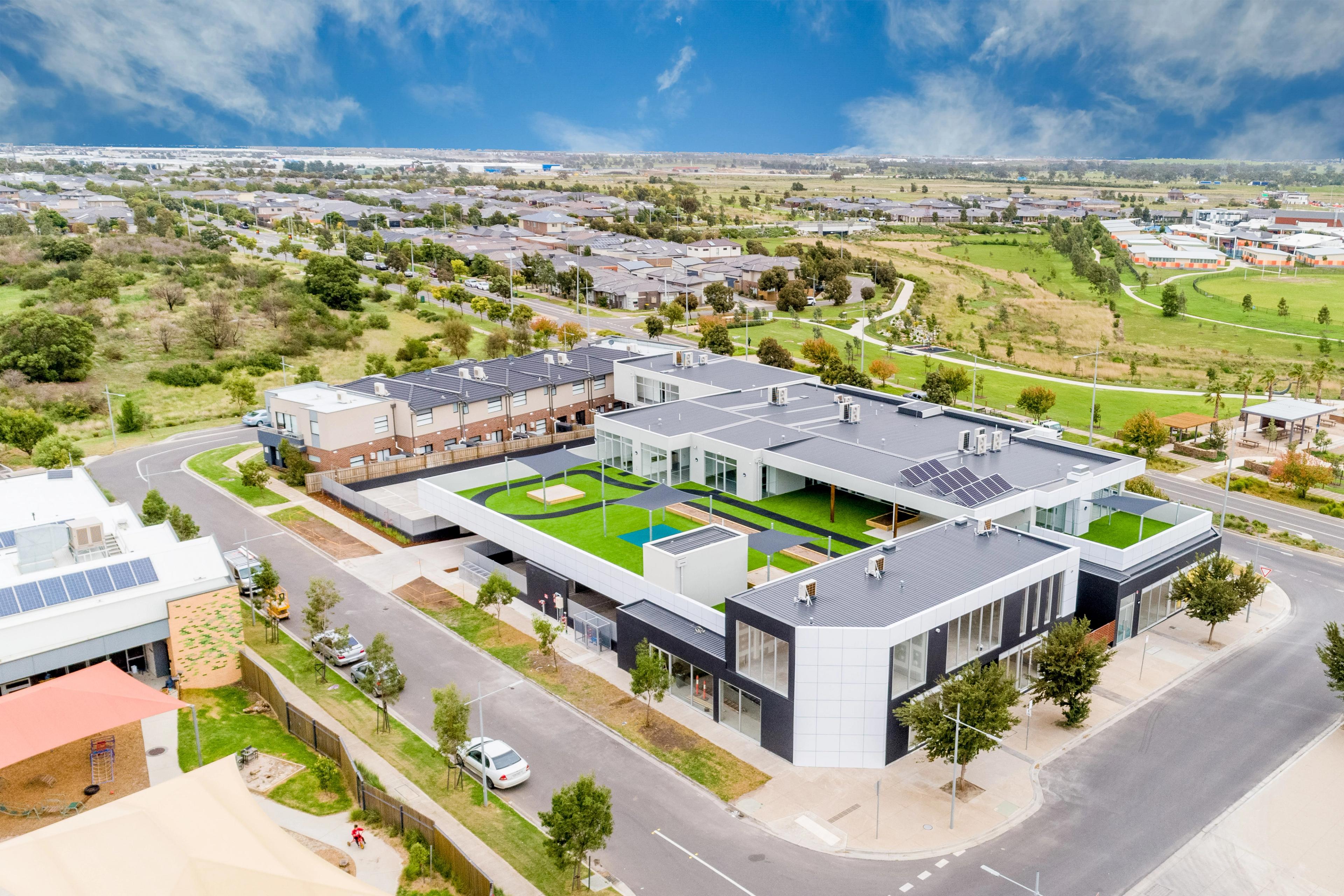 Aerial view of a modern building complex with green courtyards and solar panels, surrounded by a suburban neighborhood and green fields.