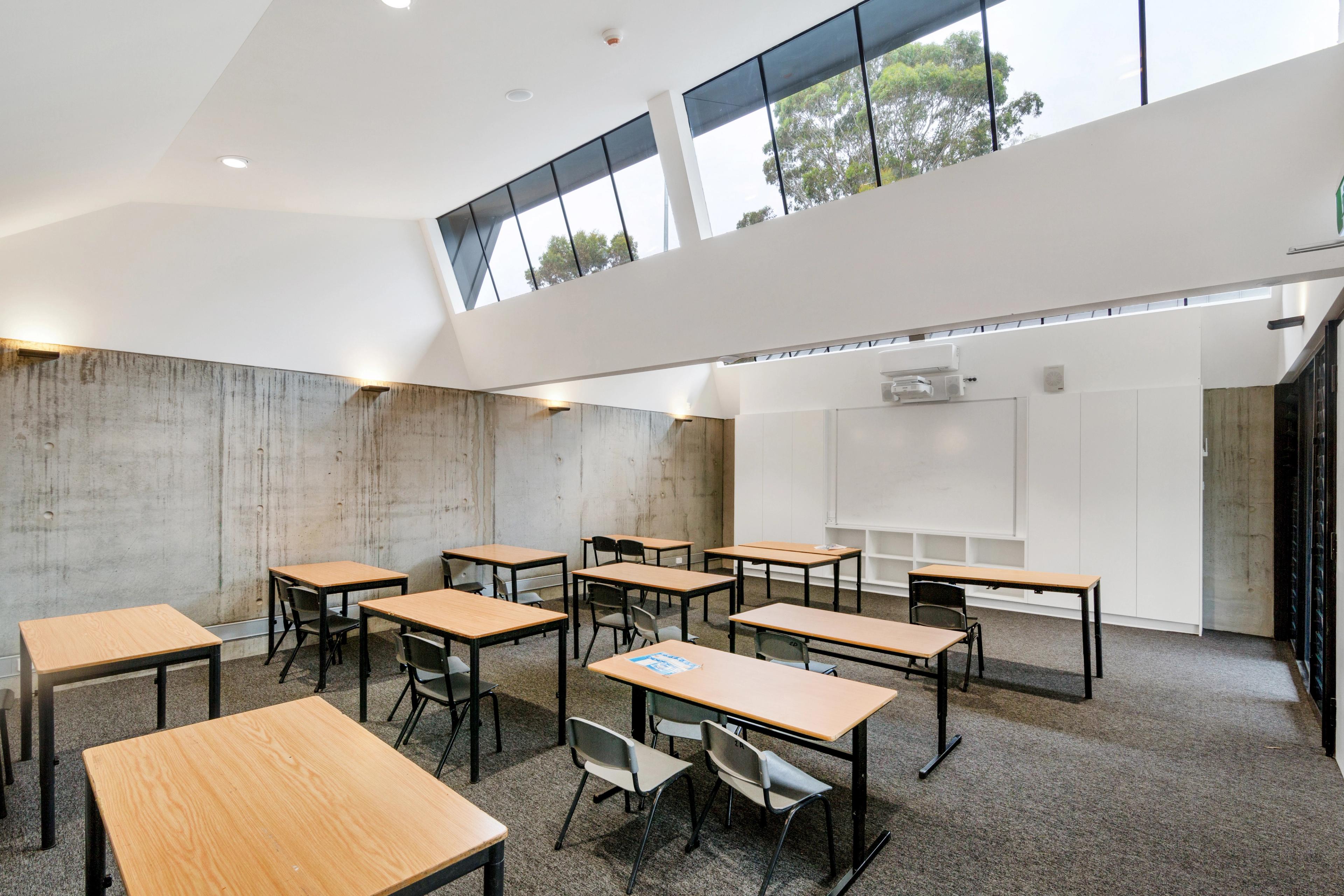 A modern classroom with individual desks and chairs, a concrete wall, high angular windows, a whiteboard, and a projector.