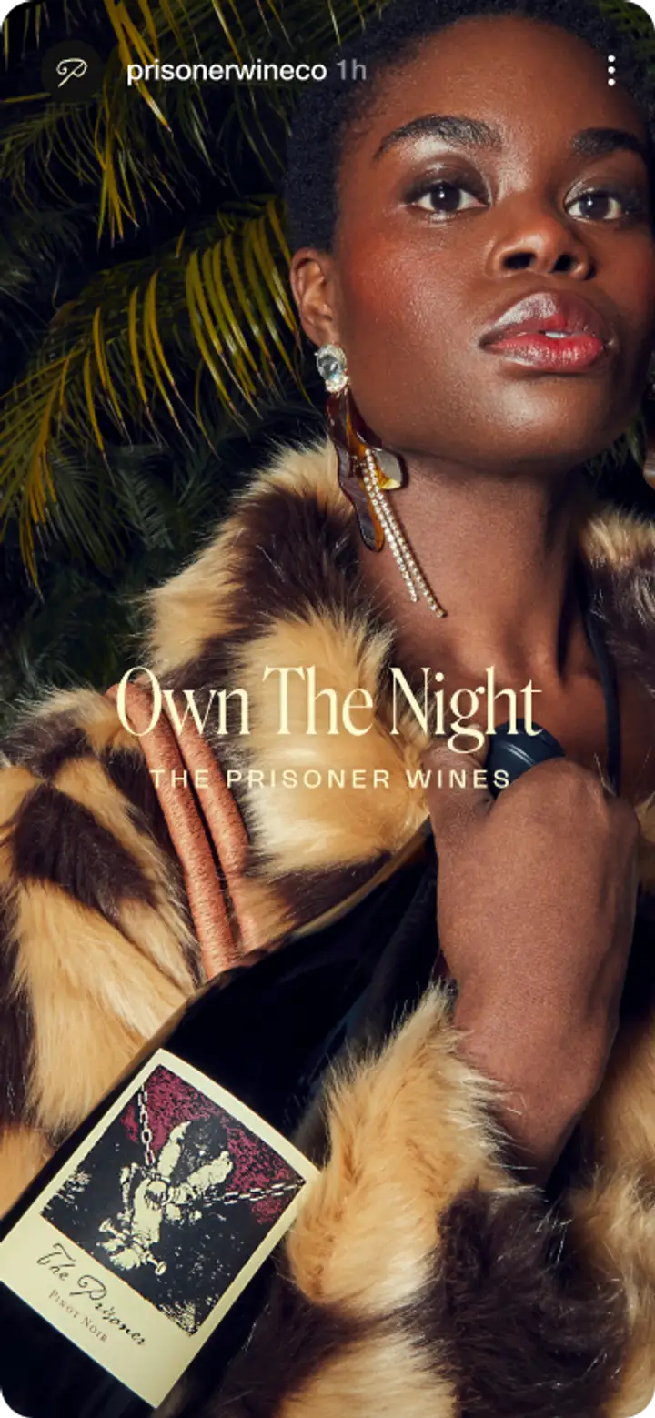 A mobile screen of a model in a fur coat holding a bottle of wine. Text says, “Own The Night, The Prisoner Wines”