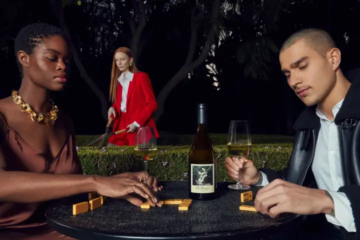 At night, two people dressed luxuriously play dominoes and hold wine glasses. A wine bottle rests on the table. In the background, a person in a bright red blazer looks at the camera while trimming a row of hedges