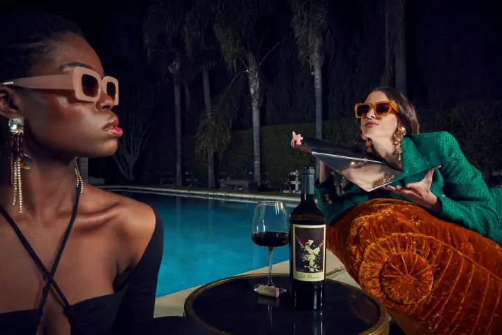 At night, two models wearing sunglasses relax next to a pool and gaze in opposite directions. One of them uses a mirror to get a tan from the moonlight