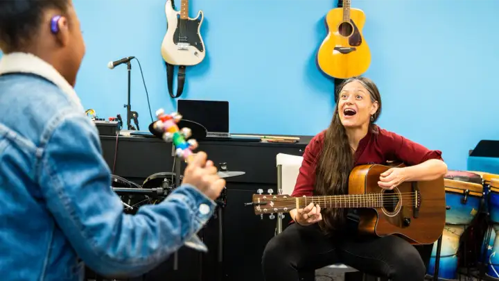 Juliana, the DMF Director of Teaching and Learning, has a huge smile while playing guitar with a music class participant who’s shaking a rainbow jingle bell