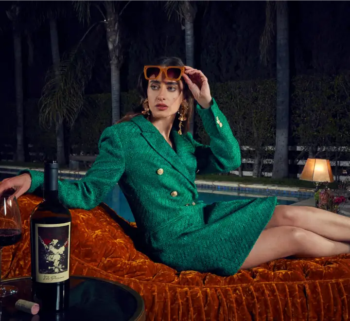 A model reclining on a couch next to a pool and a glowing lamp lifts their sunglasses to look into the distance