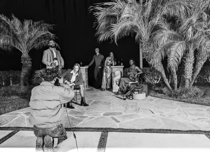 A photographer takes a photo of models posing with wine beneath palm trees at night