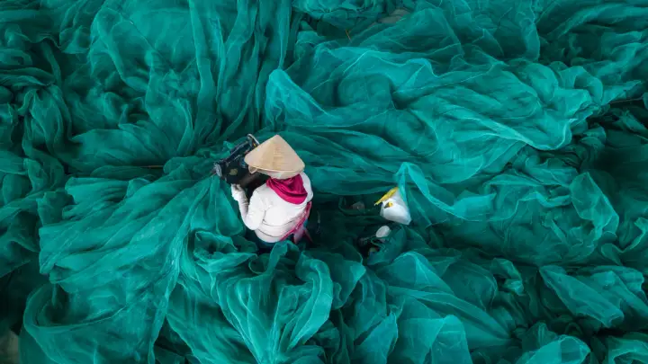 A person in a conical hat sews a fishing net from a sea of beautiful turquoise material
