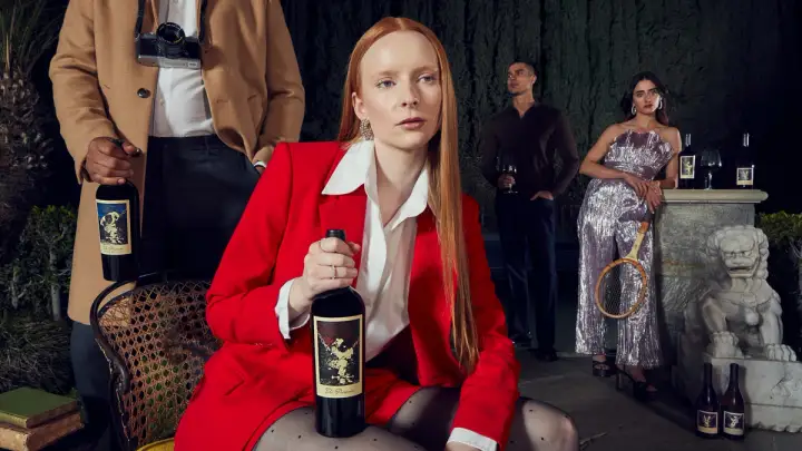A model in a bright red blazer clutches a bottle of The Prisoner Wine. In the background, another model with a camera around their neck holds a bottle of wine, a third model gazes in the distance while holding a glass of wine, and a fourth model in a shiny silver jumpsuit leans against a marble pillar while holding a racquet