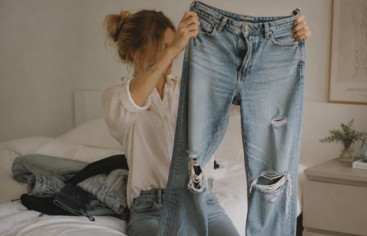 En kvinna som sitter på en säng och håller upp ett par trasiga jeans