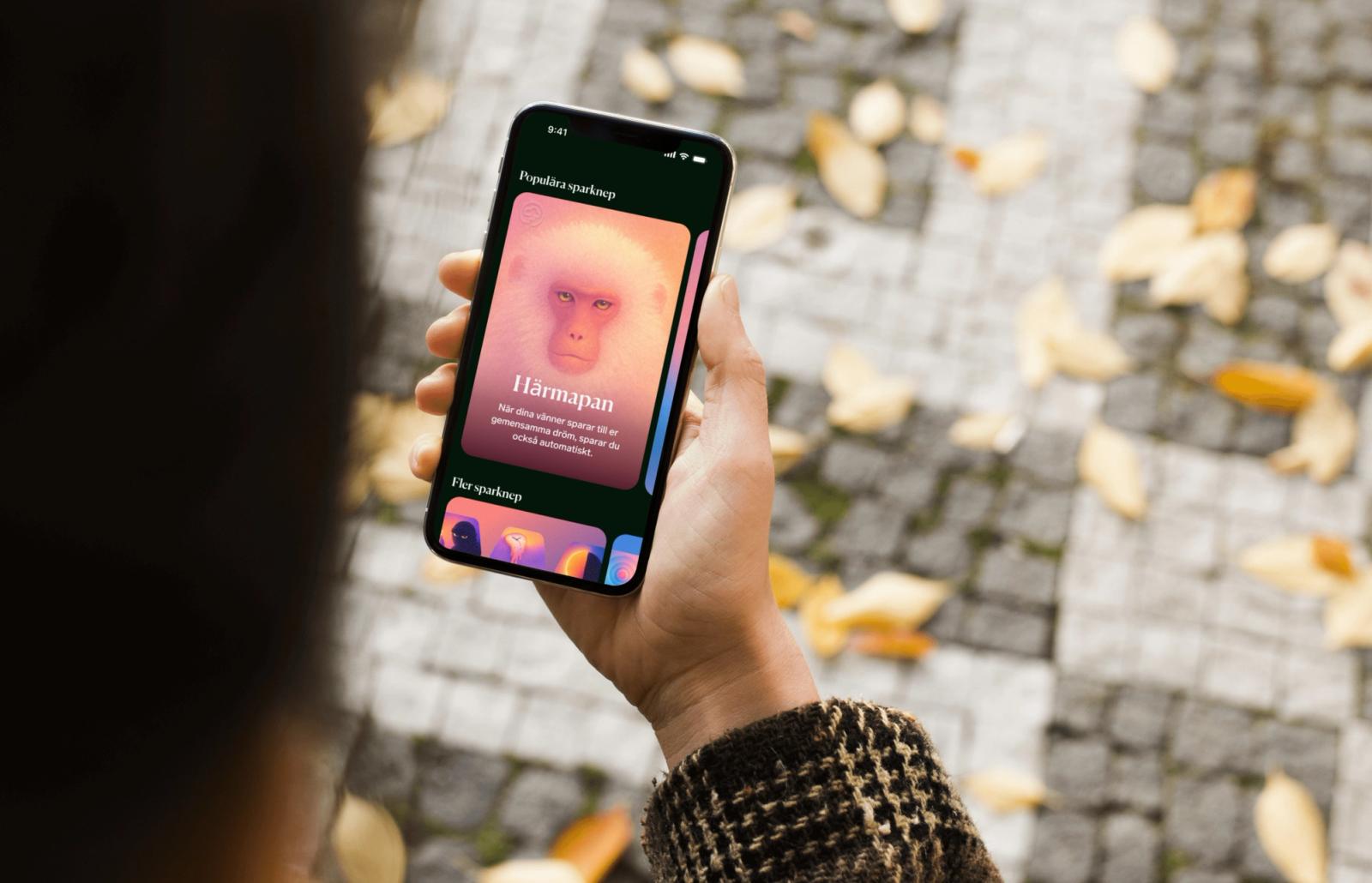 En person håller en mobiltelefon som visar sparknepet Härmapan i Dreams, med höstlöv på marken i bakgrunden.