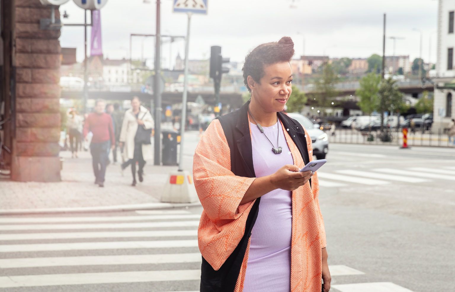 Leende person i orange omlott och lila klänning går över ett övergångsställe i centrala Stockholm med mobilen i handen