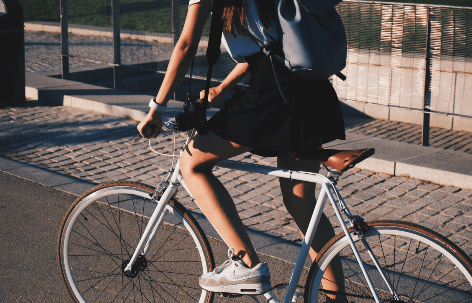 Person cyklar på vit cykel i stadsmiljö med ryggsäck på ryggen under kvällssol.