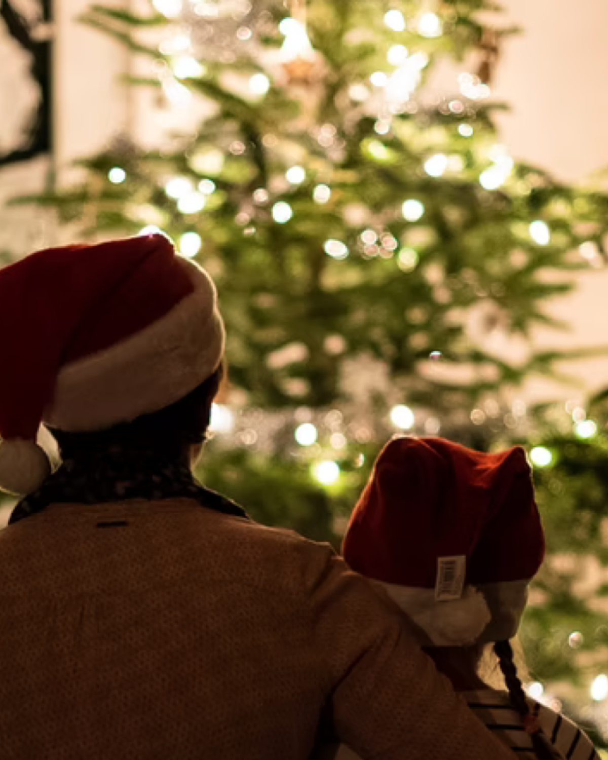Vuxen och barn med röda tomteluvor stänger av en upplyst julgran i ett mörkt rum.