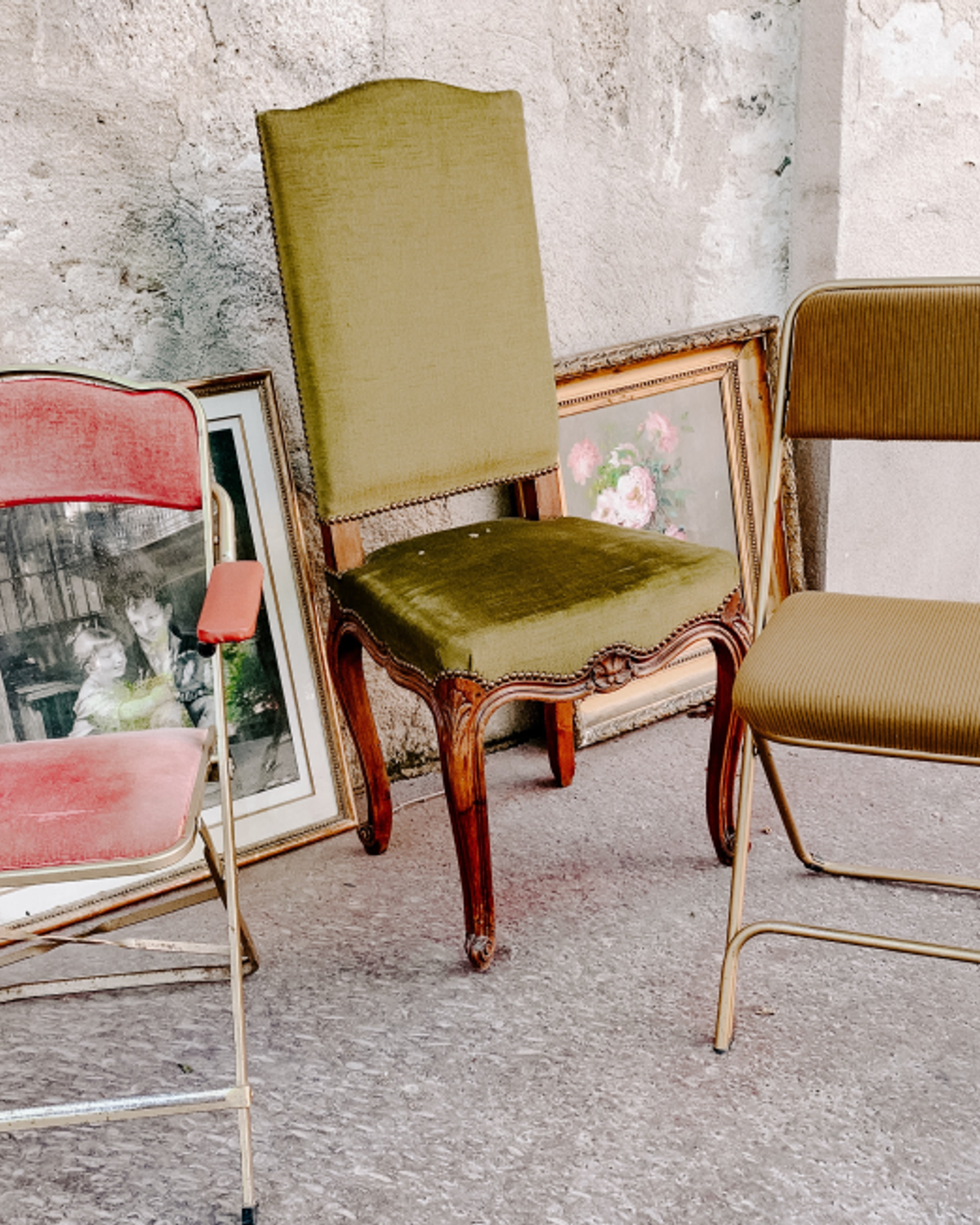 Tre begagnade stolar i olika färger uppställda mot en gammal stenvägg — vintageinredning på loppmarknad