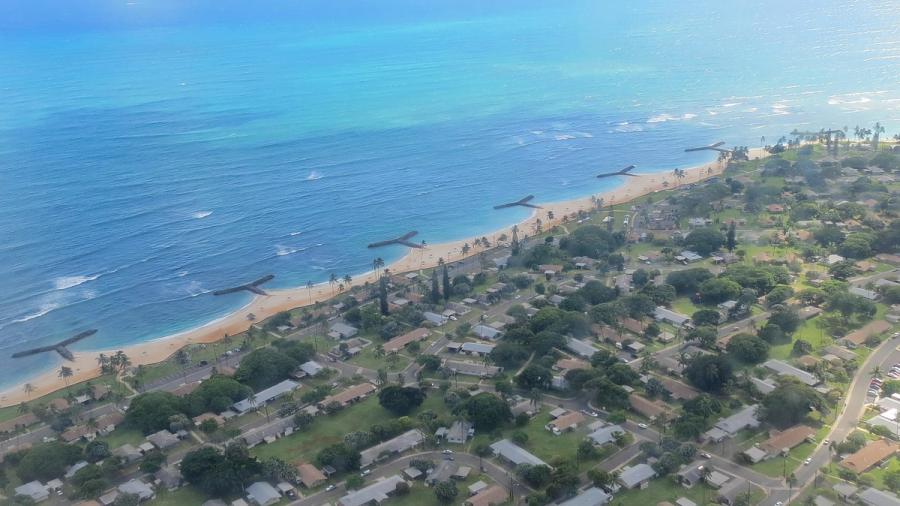 Aerial view of ʻEwa Beach on Oʻahu, showing a long sandy shoreline with rock groynes, palm trees, and neighborhoods along the coast.