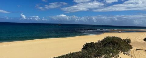 Vast expanse of golden sand stretching along Polihale State Park with deep blue ocean and distant sailboat