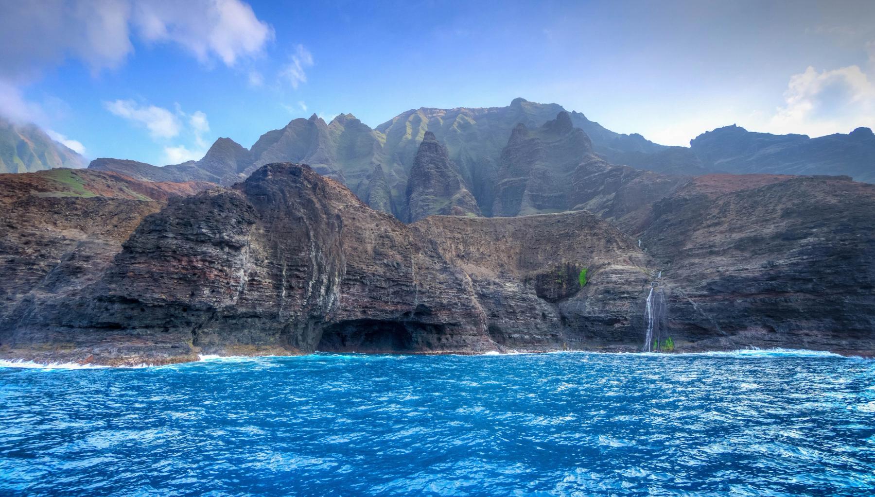 Offshore view of Nā Pali Coast cliffs with sea caves and thin waterfalls above bright blue ocean under a clear sky