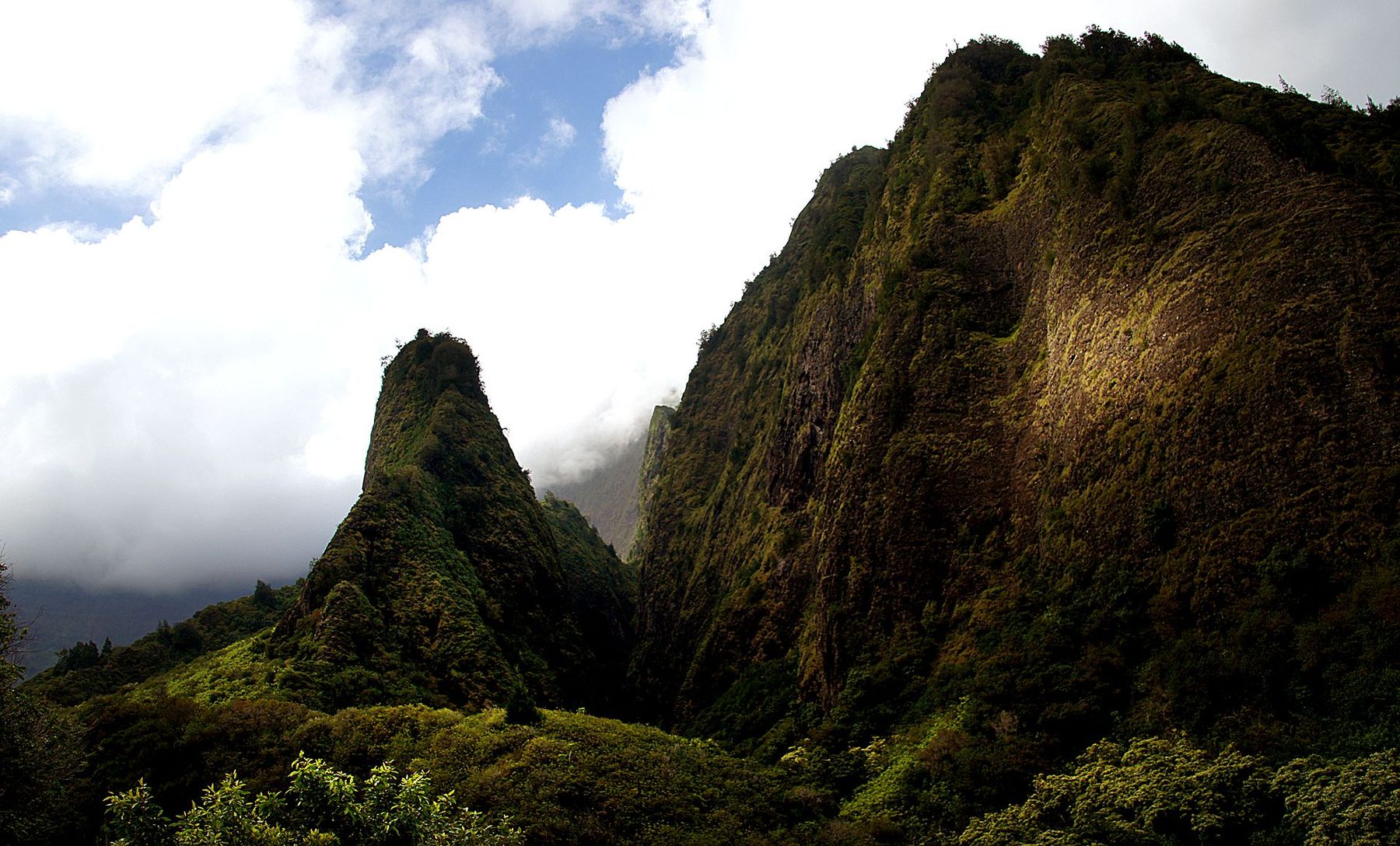 Steep, green mountain ridges rise above dense forest with low clouds and patches of blue sky in ʻĪao Valley State Park on Maui.