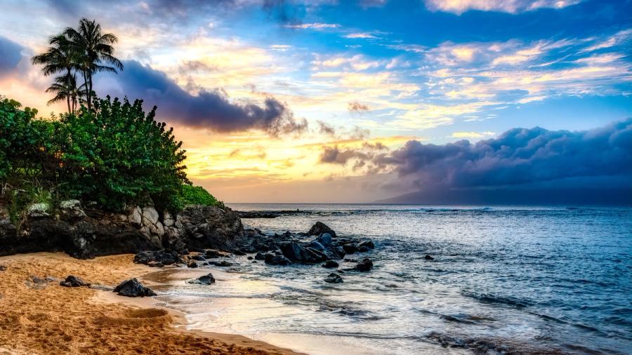 Rocky shoreline and small sandy beach with palm trees at sunset, with waves under dramatic clouds in Kapalua, Maui.