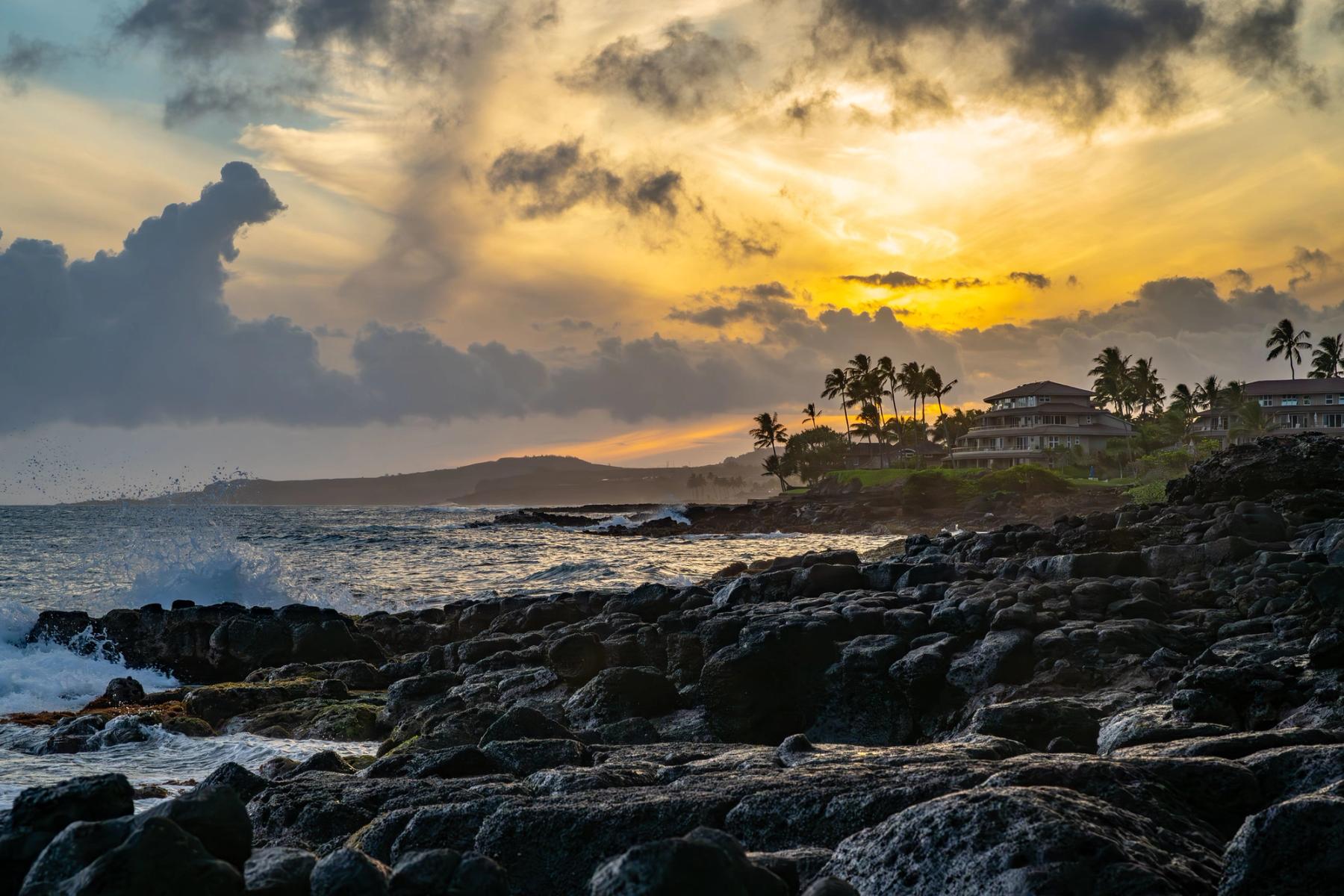 Golden sunset over the ocean from a rocky lava shoreline, with wave spray, dramatic clouds, and palm trees beside beachfront buildings in Poʻipū