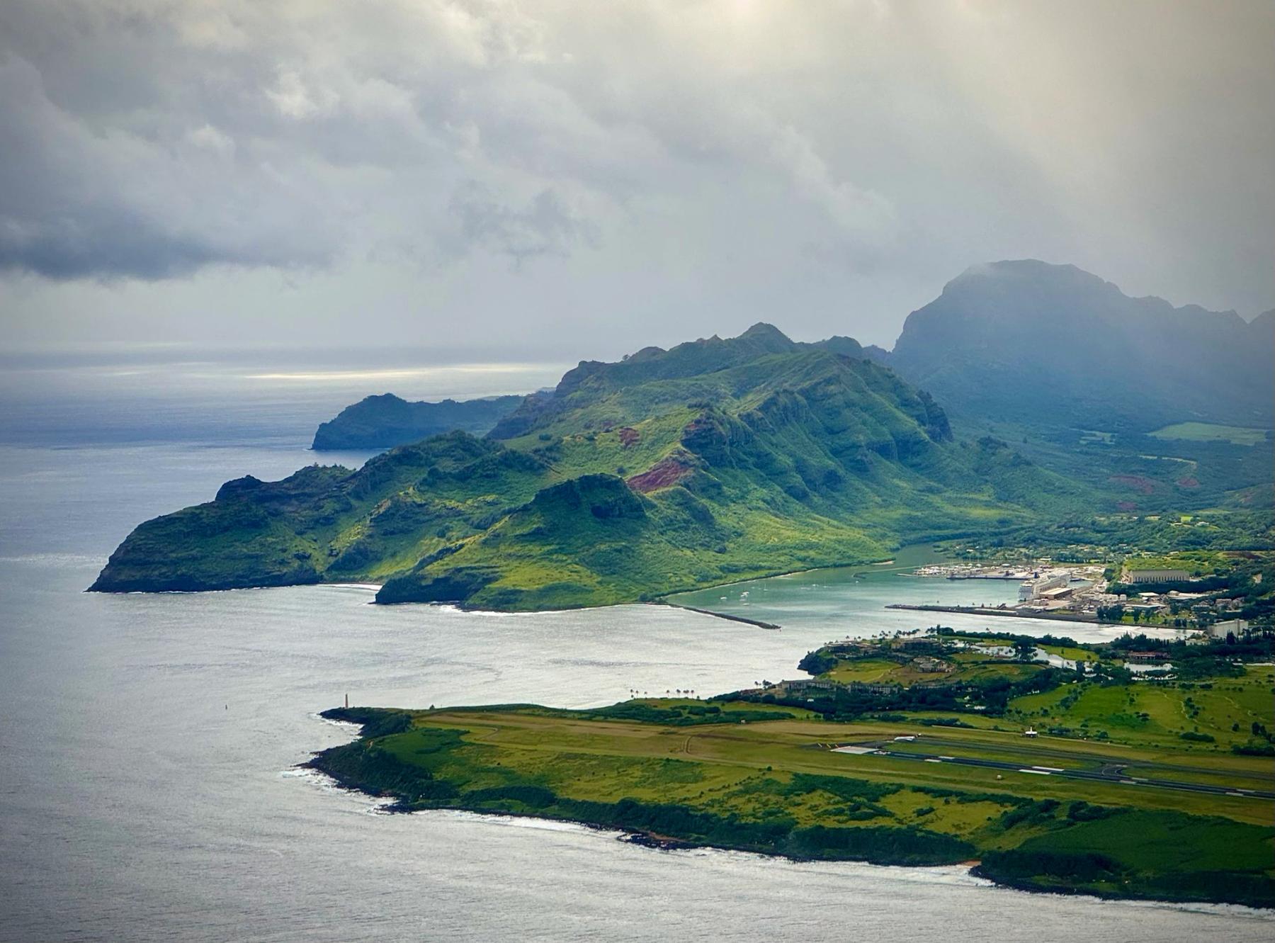 Aerial view of Kauaʻi with green mountain ridges, curving coastline, a harbor, and a runway under hazy sunlight