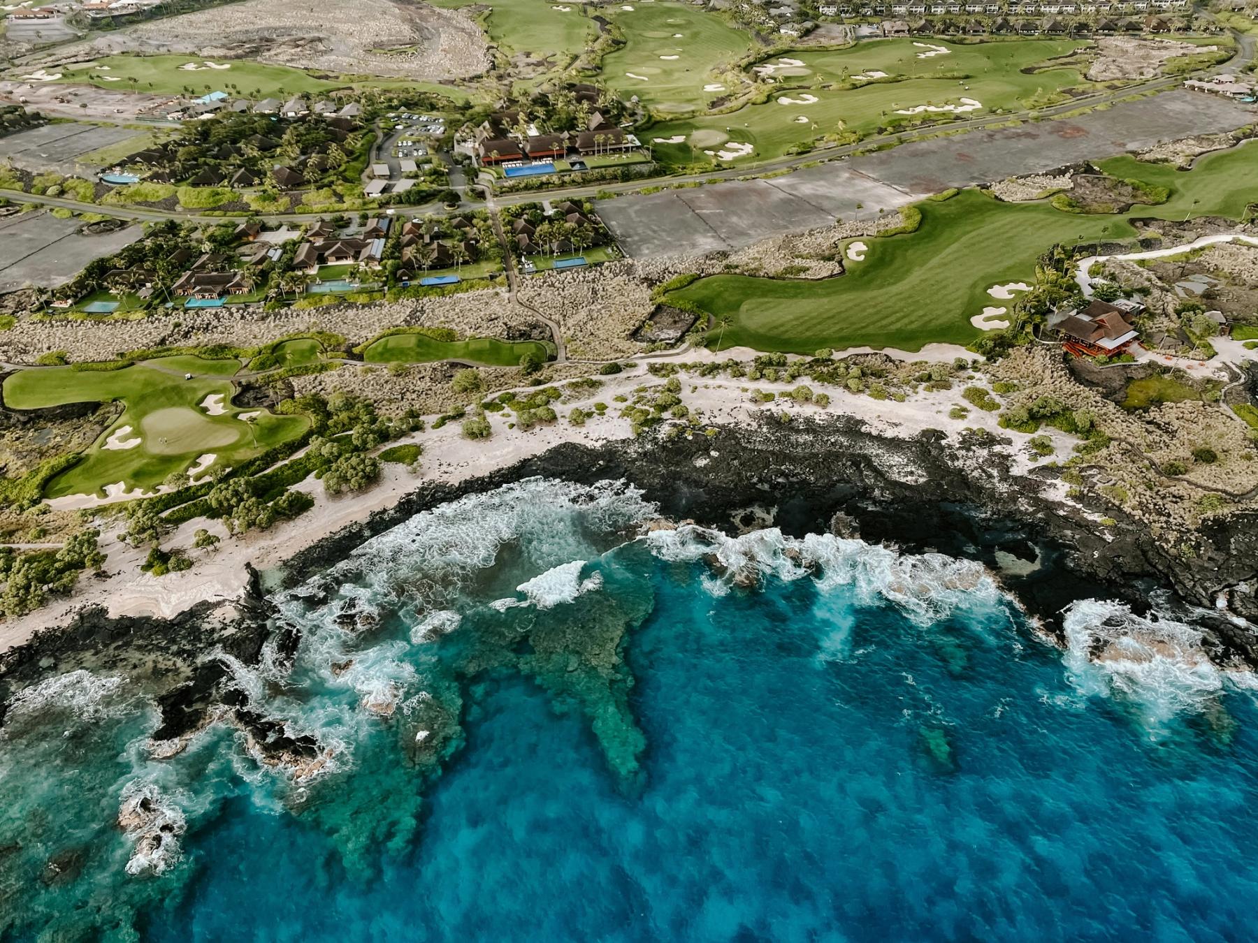 Aerial view of Kona’s rocky shoreline with turquoise surf, a narrow beach, and green golf course fairways and buildings inland.