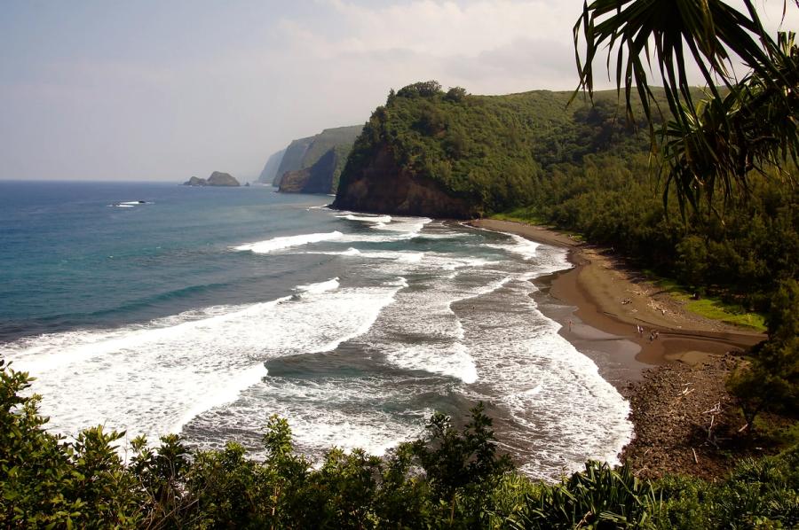 Waves roll onto a dark sand beach beneath steep green sea cliffs at Pololū Beach in North Kohala on Hawaiʻi Island.