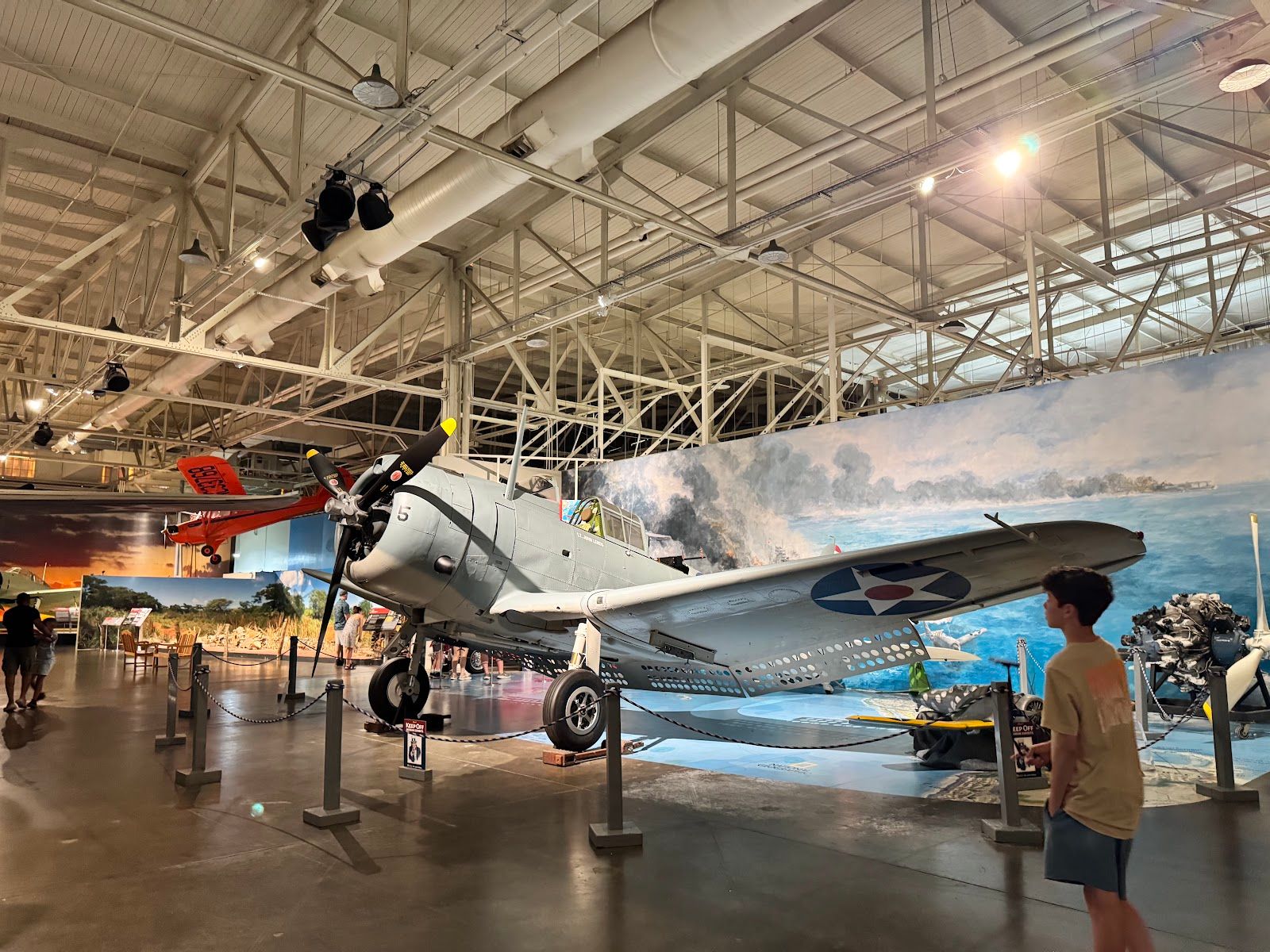 An indoor aircraft exhibit at Pearl Harbor Aviation Museum with visitors walking through the hangar gallery.