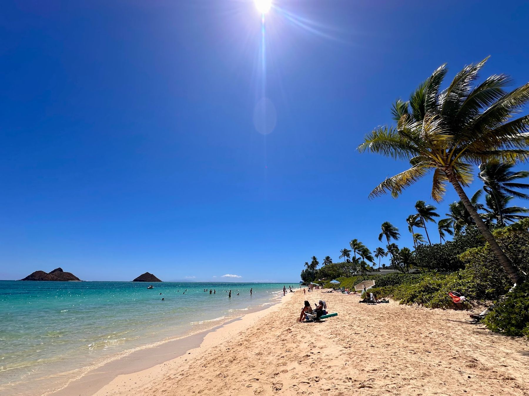 Beachgoers on the white sand shoreline at Lanikai Beach with palms, blue water, and the Mokulua Islands offshore.