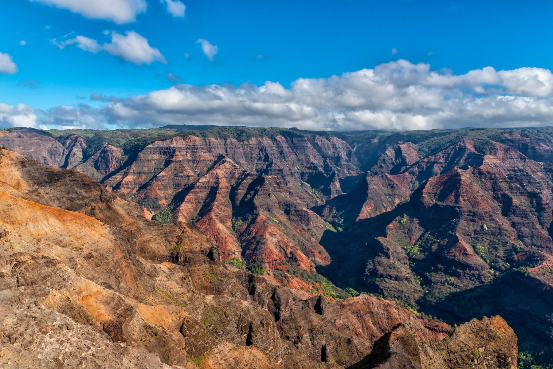 Wide view across Waimea Canyon with red stratified ridges, deep shadowed valleys, and scattered clouds under a bright blue sky