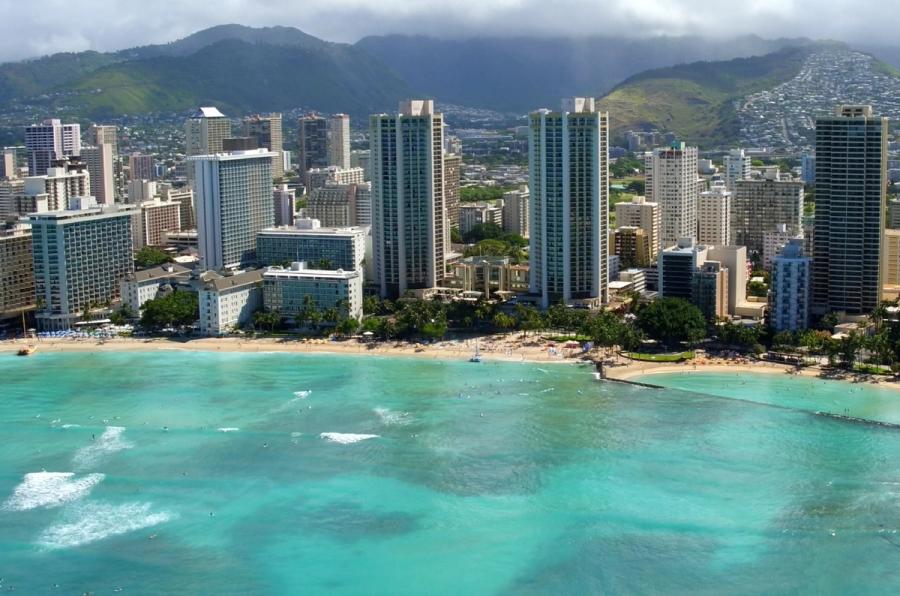 Aerial view of turquoise ocean along a sandy beach lined with palm trees and high-rise hotels, with green mountains in the background in Waikīkī.