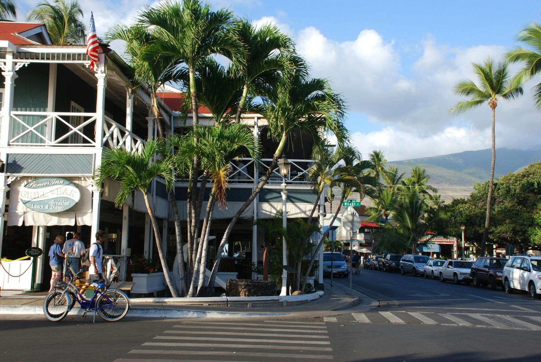 Street corner in Lāhainā, Maui with palm trees, a white building, parked cars, pedestrians, and bicycles near a crosswalk.