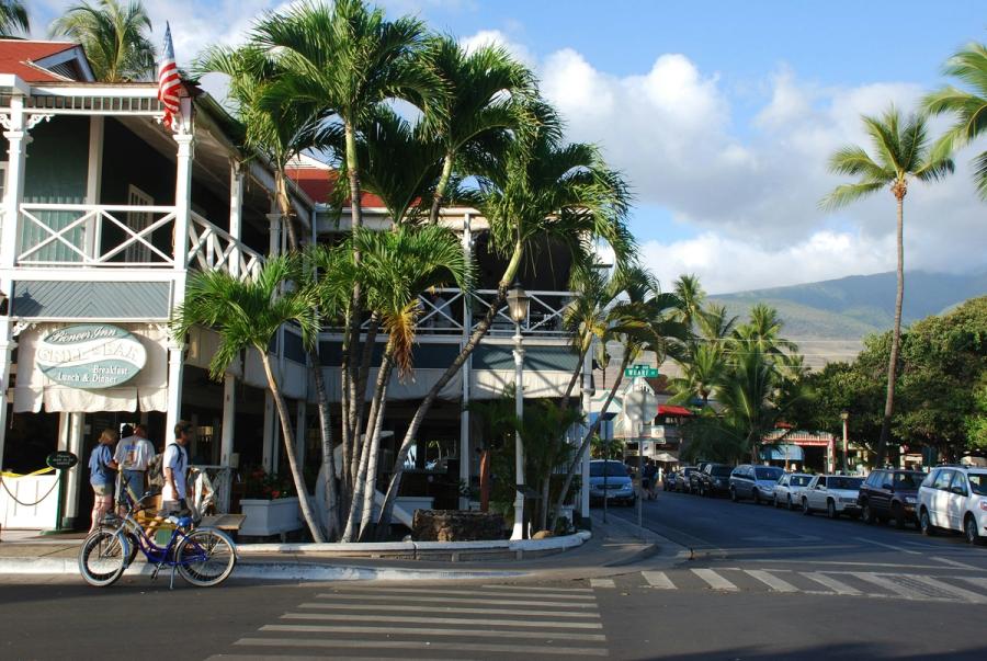 Street corner in Lāhainā, Maui with palm trees, a white building, parked cars, pedestrians, and bicycles near a crosswalk.