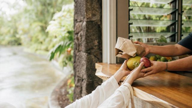 Hands pass a paper bag and fruit through a wooden service window beside louvered glass, with greenery in the background.