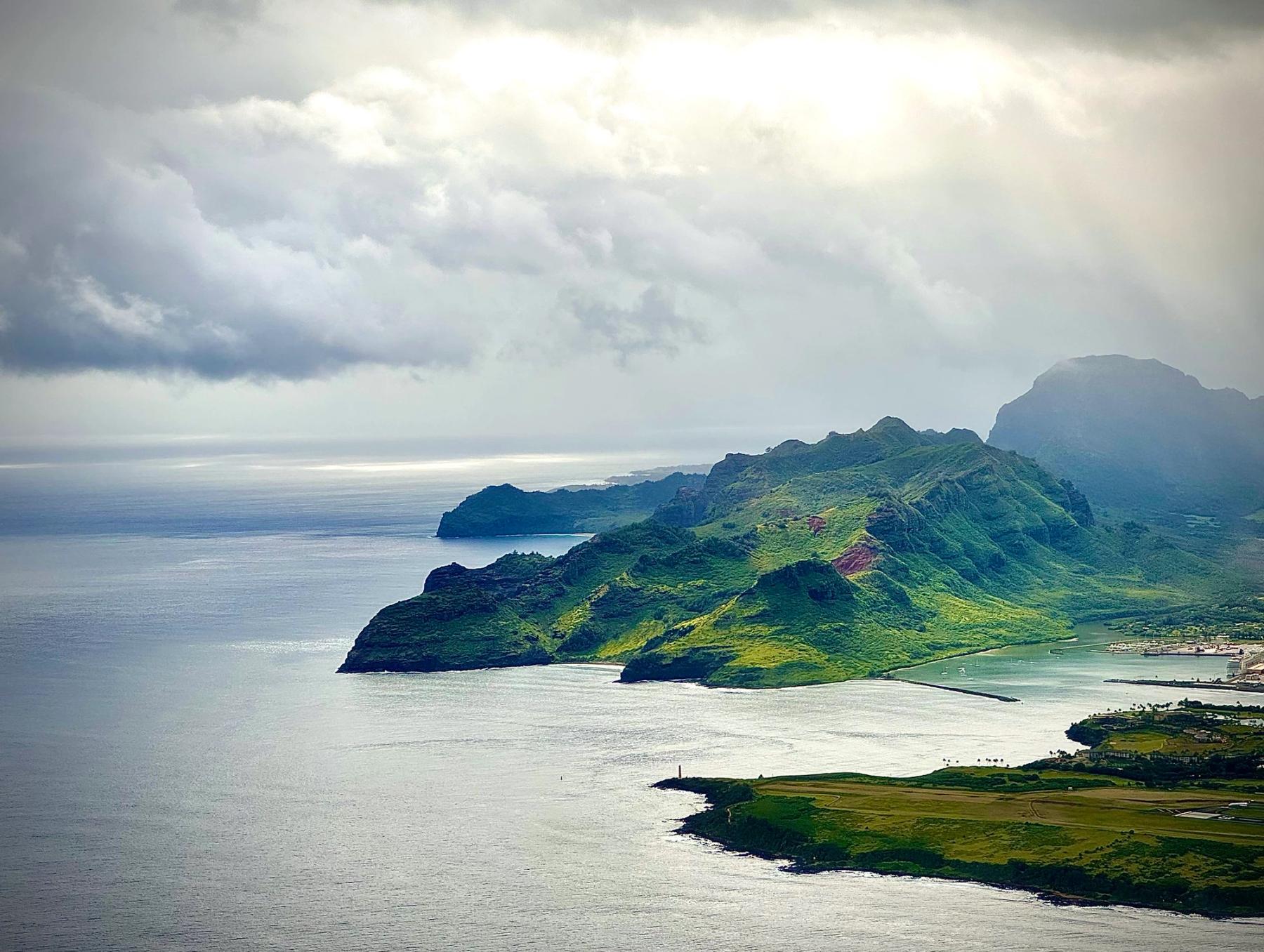 Aerial view of Kauaʻi’s lush green headlands and Nawiliwili Bay under dramatic clouds with bright sun breaking through over the ocean.